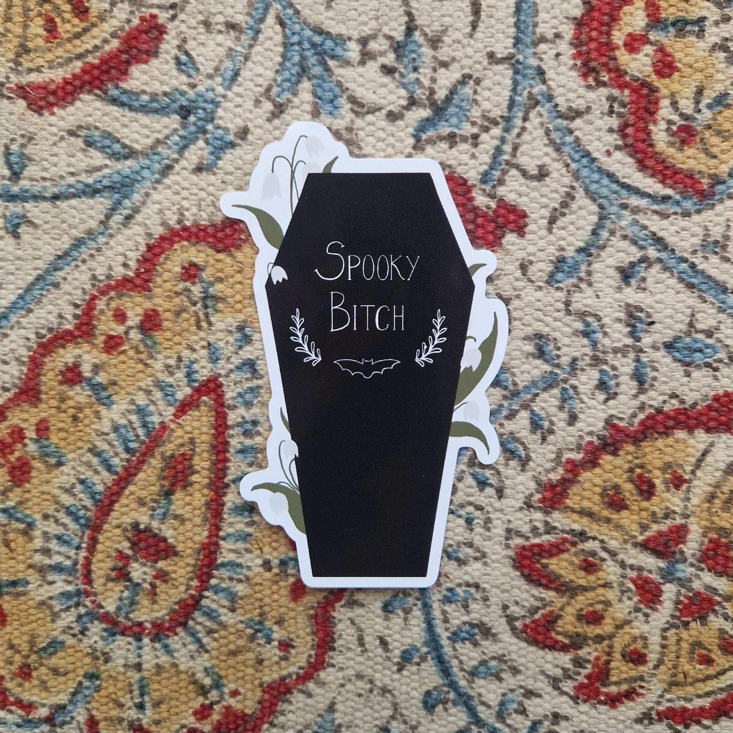 A black coffin sticker with the words 'SPOOKY BITCH' in white across the middle with a bat silhoette and leafy vines design underneath. Around the edges of the coffin are lily of the valley flowering plants. Behind the sticker is a cream fabric with a red, yellow and blue pattern.