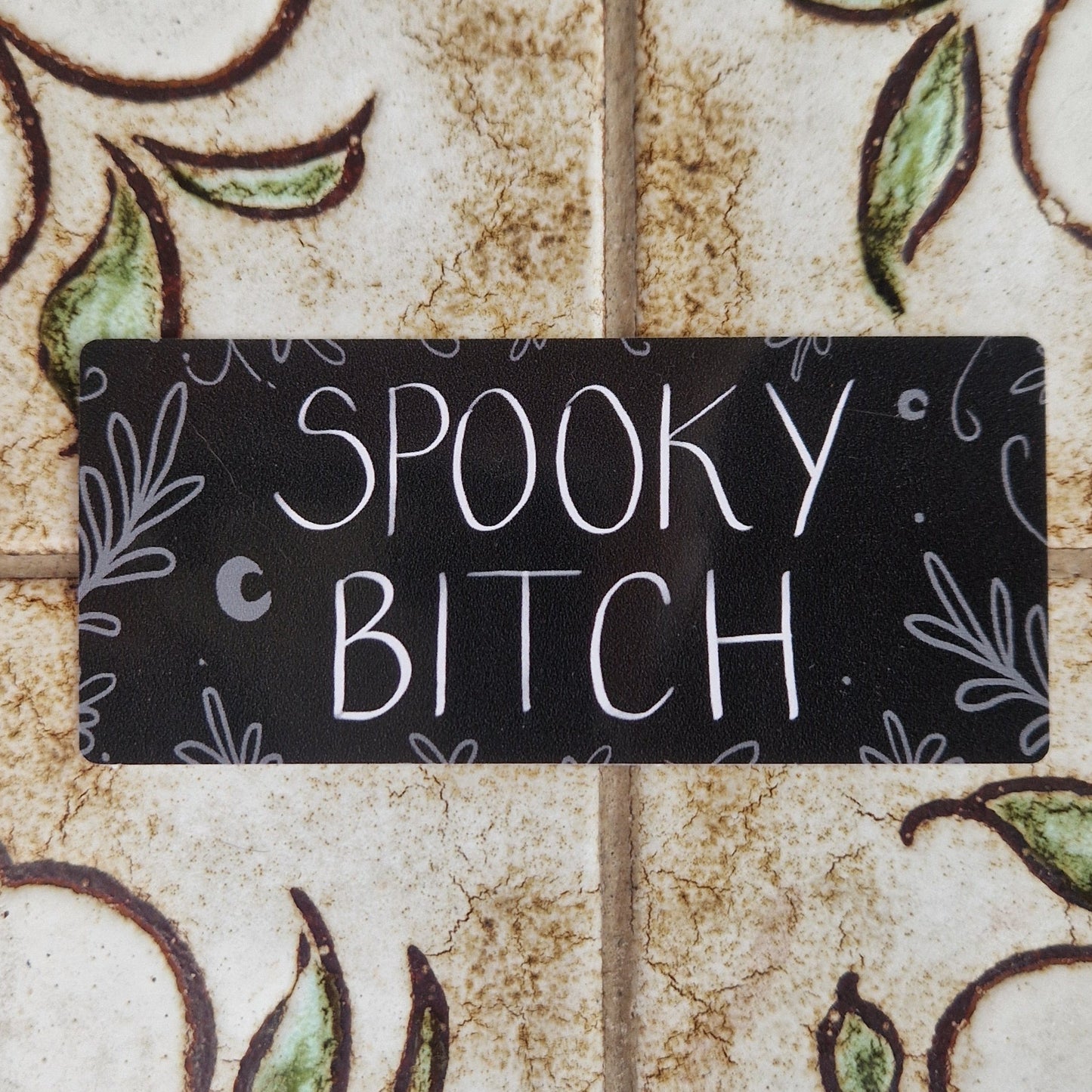 Rectangular black vinyl sticker with the words 'Spooky Bitch' across the middle in black writing and grey leafy vines around the edges and a couple of crescent moons.