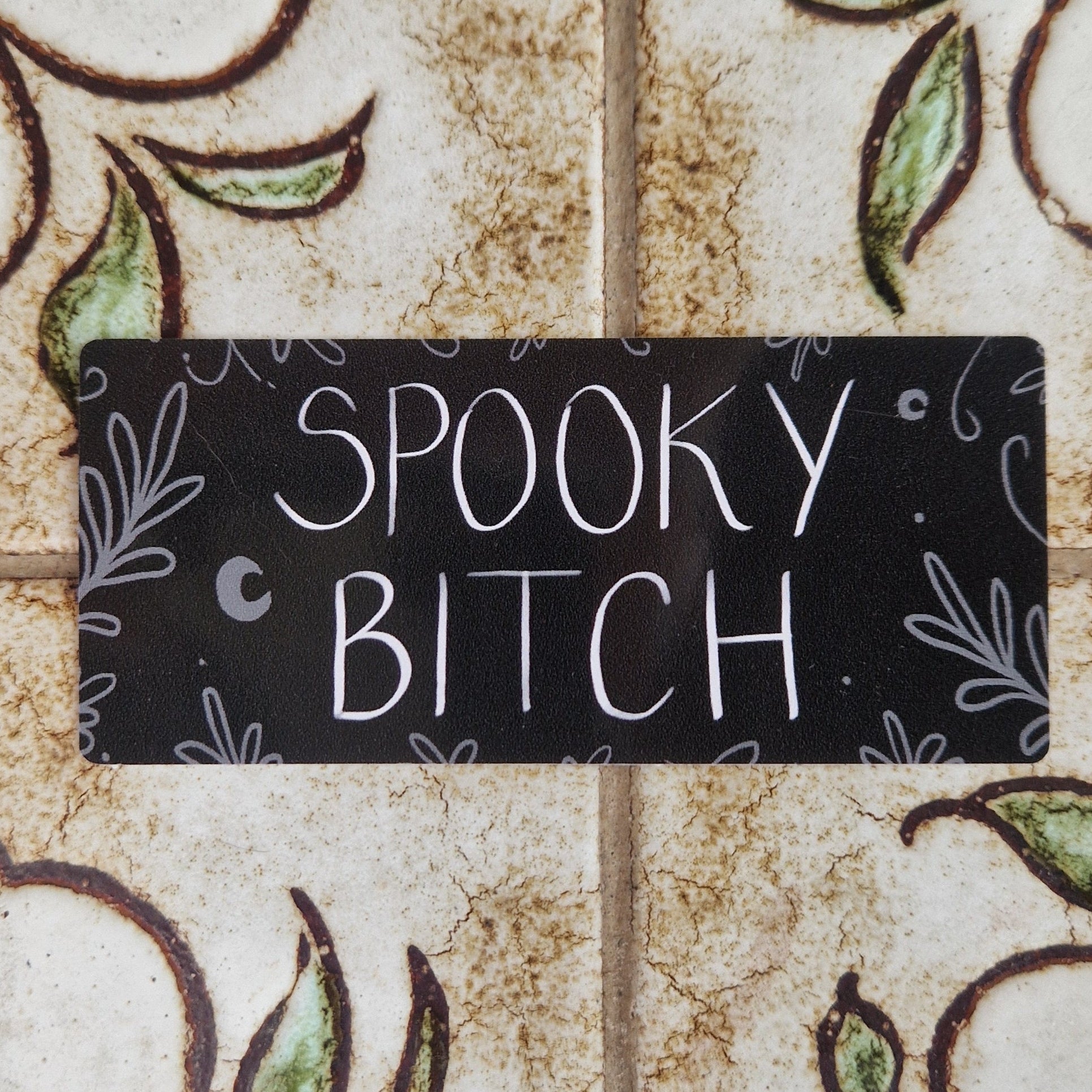 Rectangular black vinyl sticker with the words 'Spooky Bitch' across the middle in black writing and grey leafy vines around the edges and a couple of crescent moons.