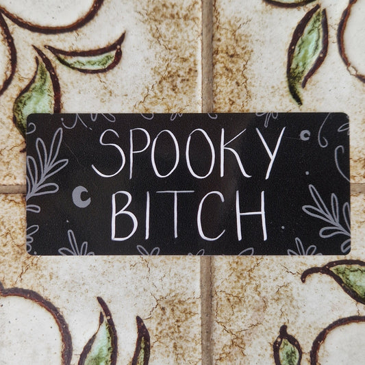 Rectangular black vinyl sticker with the words 'Spooky Bitch' across the middle in black writing and grey leafy vines around the edges and a couple of crescent moons.