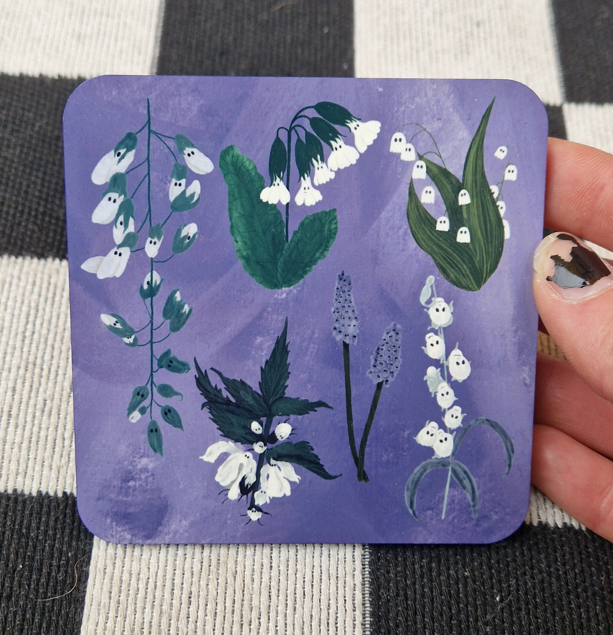 A square coaster with a mottled purple background is held by a white hand in front of a checkered white and black fabric. The coaster features six different plants with ghosts instead of their usual flowers.