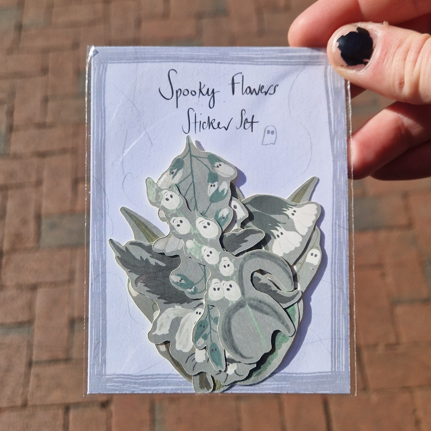 A white hand with chipped black nail polish holds a Spooky Flowers Sticker Set in front of some orange paving slabs. You can see different ghost themed flower stickers with grey backgrounds inside the pack.