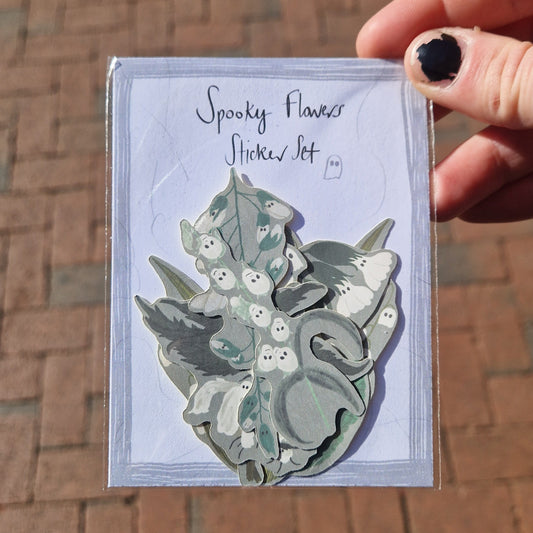 A white hand with chipped black nail polish holds a Spooky Flowers Sticker Set in front of some orange paving slabs. You can see different ghost themed flower stickers with grey backgrounds inside the pack.