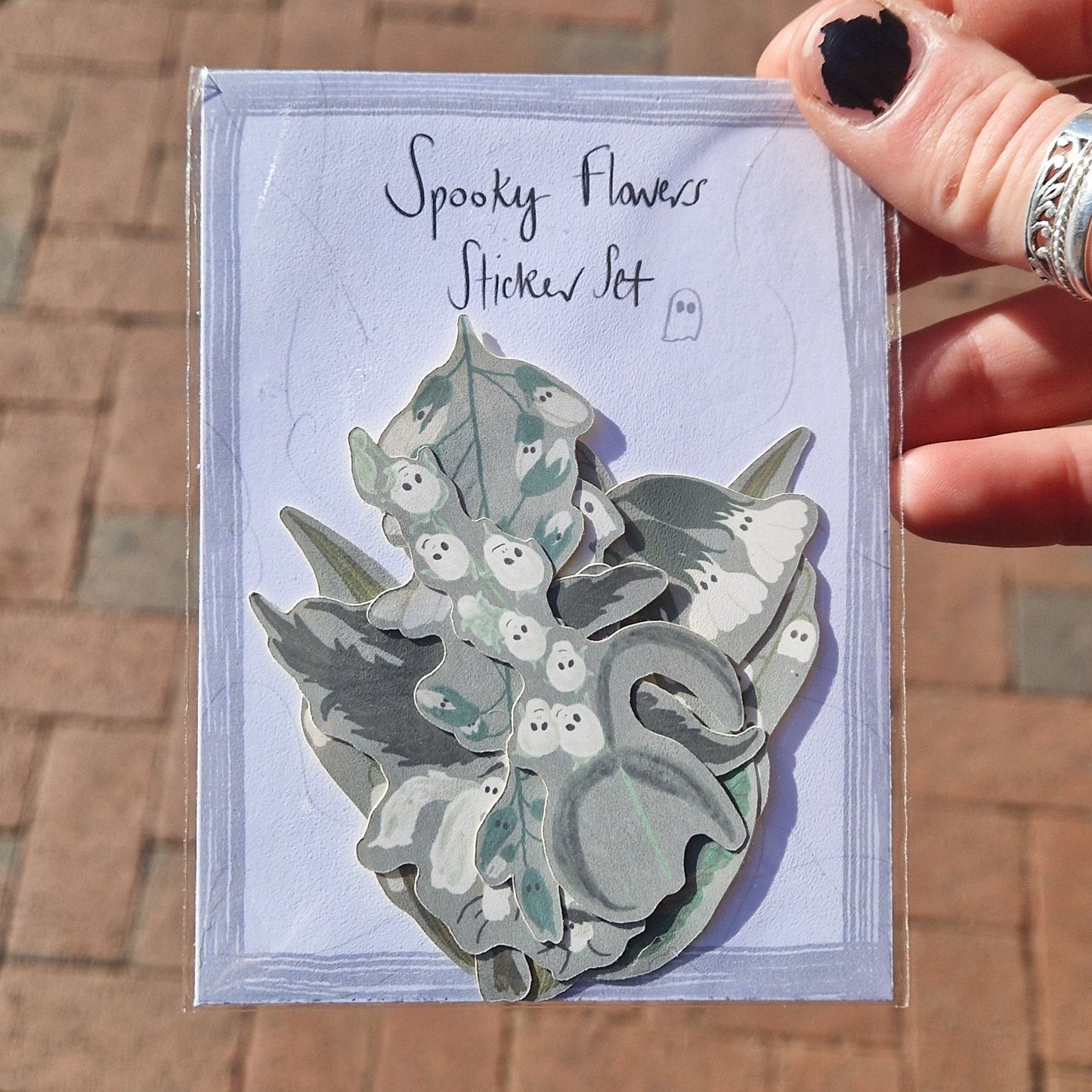 A white hand with chipped black nail polish holds a Spooky Flowers Sticker Set in front of some orange paving slabs. You can see different ghost themed flower stickers with grey backgrounds inside the pack.