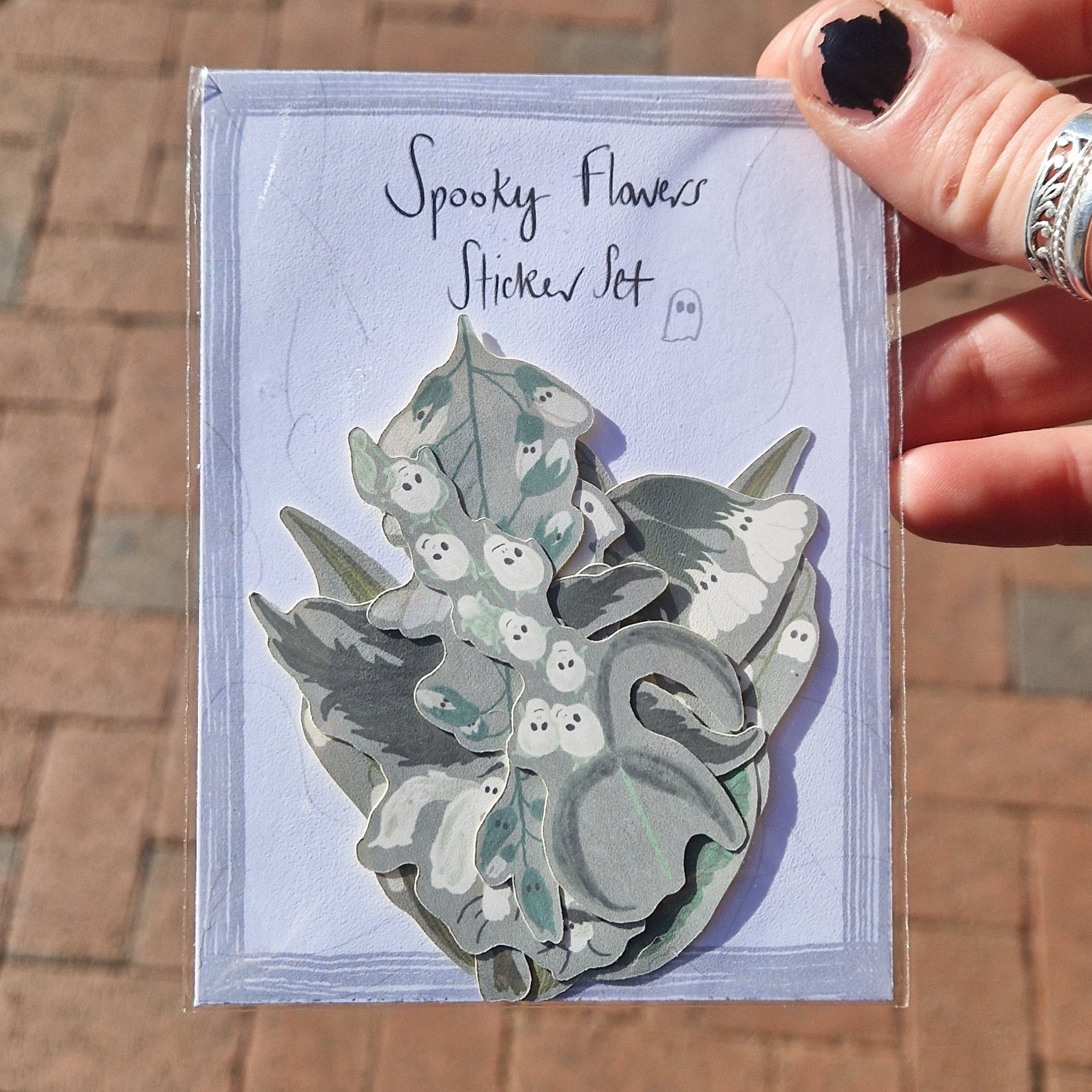 A white hand with chipped black nail polish holds a Spooky Flowers Sticker Set in front of some orange paving slabs. You can see different ghost themed flower stickers with grey backgrounds inside the pack.