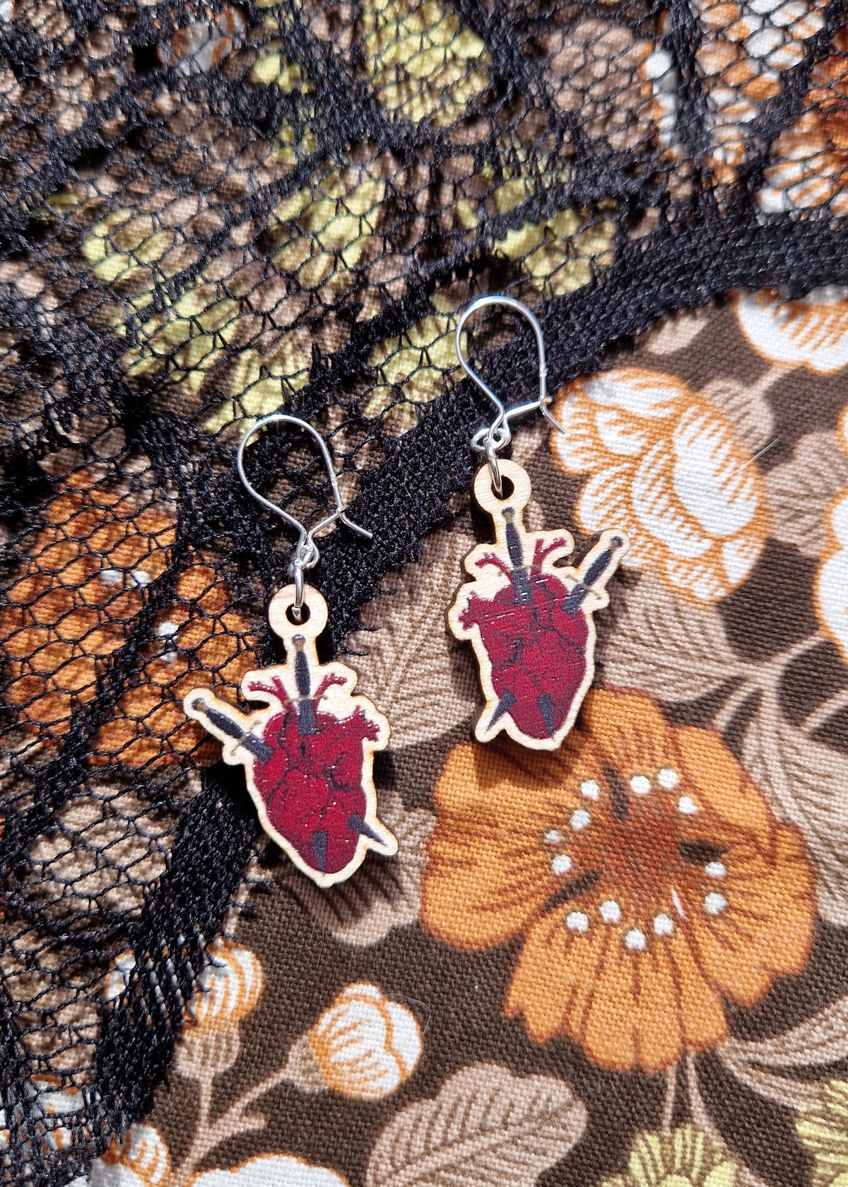 A pair of red anatomical heart and dagger earrings with two daggers through each heart, sit on a black lace and floral fabric background. The earrings have silver jump rings and ear wires.