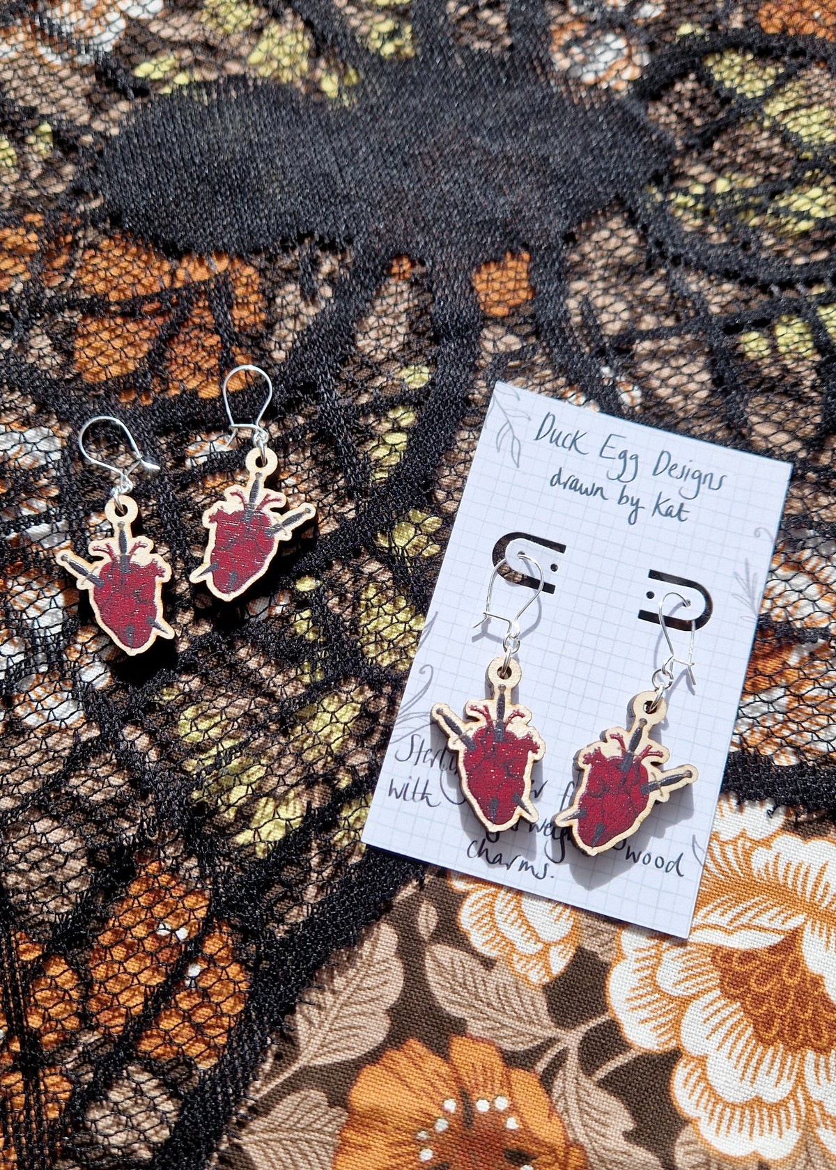 Two pairs of anatomical heart and dagger earrings sit next to each other on a black lace and floral fabric background. The pair on the right are on a white backing card that has the words 'Duck Egg Designs drawn by Kat' across the top.