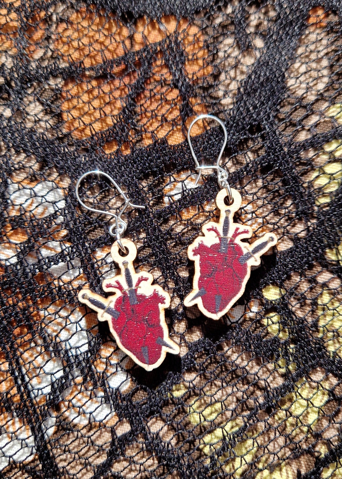 A pair of red, black and grey  anatomical heart earrings with two daggers through them - one from top to bottom and one from left to right (or right to left). The earrings have sterling silver jump rings and ear wires.