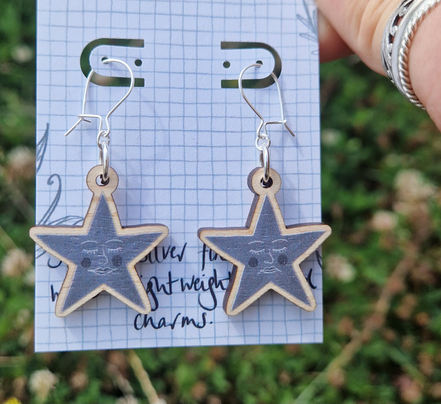 A pair of grey star earrings featuring one happy face and one sad face, hang from a white backing card with a grey grid design. The backing card is held in front of a green leafy garden by a white hand. 