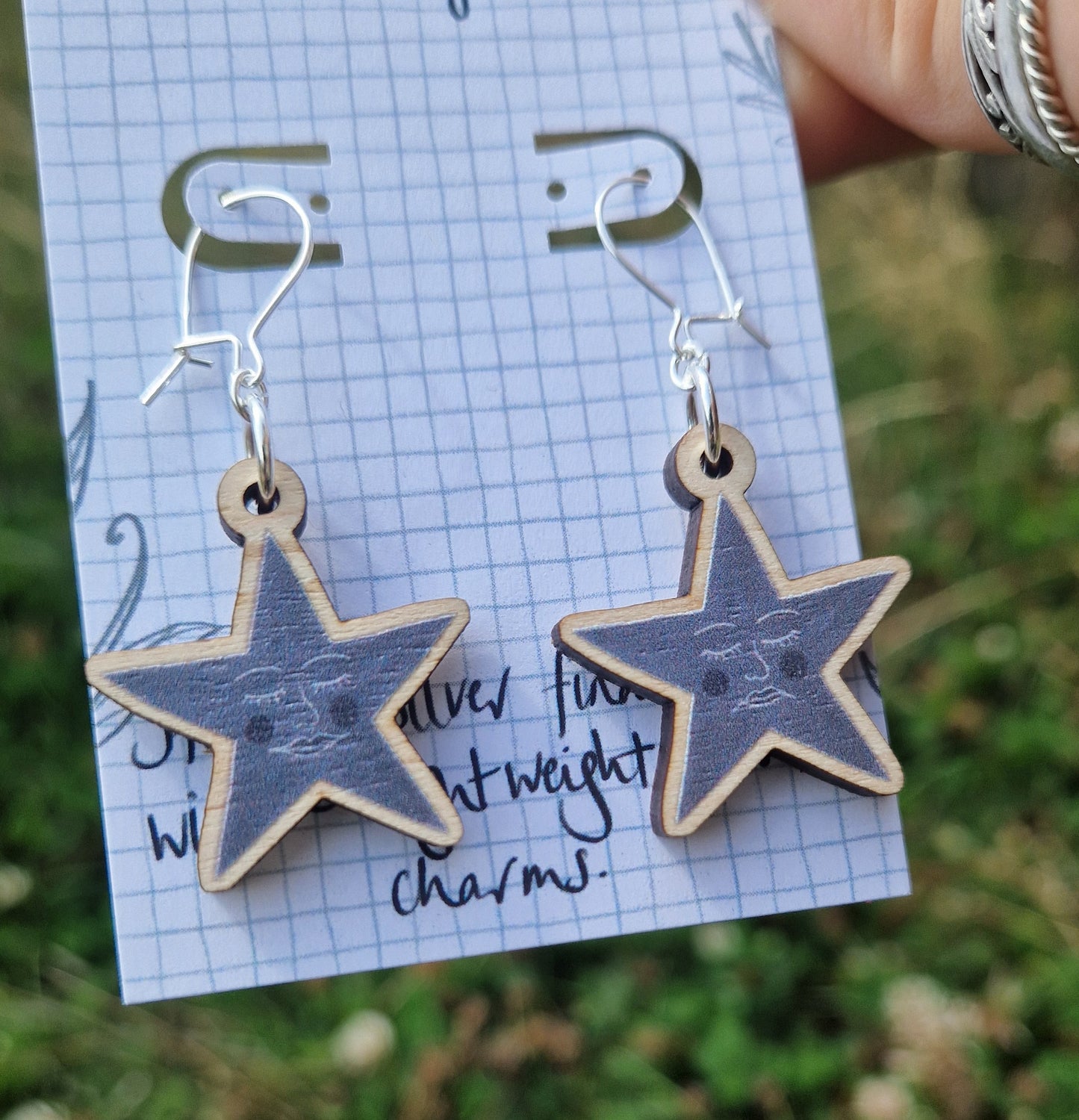 A pair of grey star earrings featuring one happy face and one sad face, hang from a white backing card. . The backing card is held in front of a green leafy garden by a white hand with silver rings.