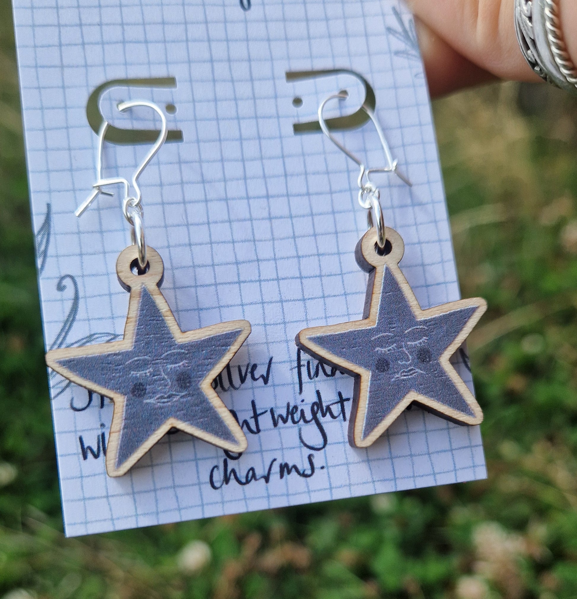 A pair of grey star earrings featuring one happy face and one sad face, hang from a white backing card. . The backing card is held in front of a green leafy garden by a white hand with silver rings.