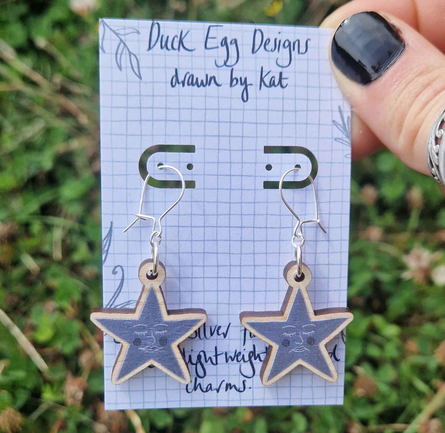 A pair of grey star earrings featuring one happy face and one sad face, hang from a white backing card with a grey grid design and the words 'Duck Egg Designs, drawn by Kat' across the top in black handwriting. The backing card is held in front of a green leafy garden by a white hand with black nail polish.