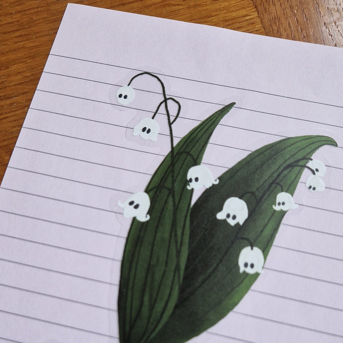 Close up image of the ghost flowers on the lily of the valley plant sticker. The sticker has a transparent background and is on a lined lilac sheet of paper.