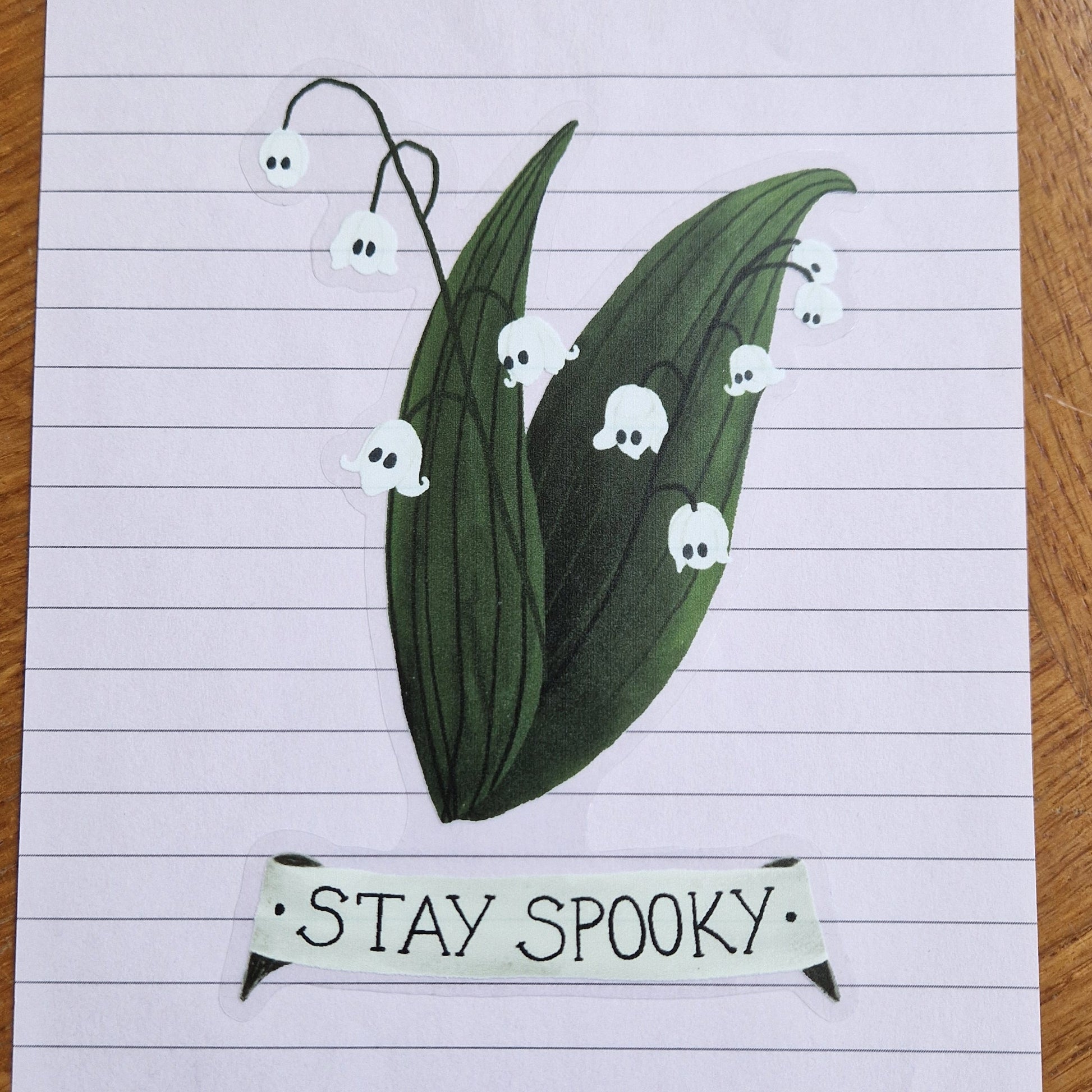 A lined lilac sheet of paper with a sticker featuring a lily of the valley plant with ghosts for flowers and a banner underneath that reads 'STAY SPOOKY'. The sticker has a transparent background.