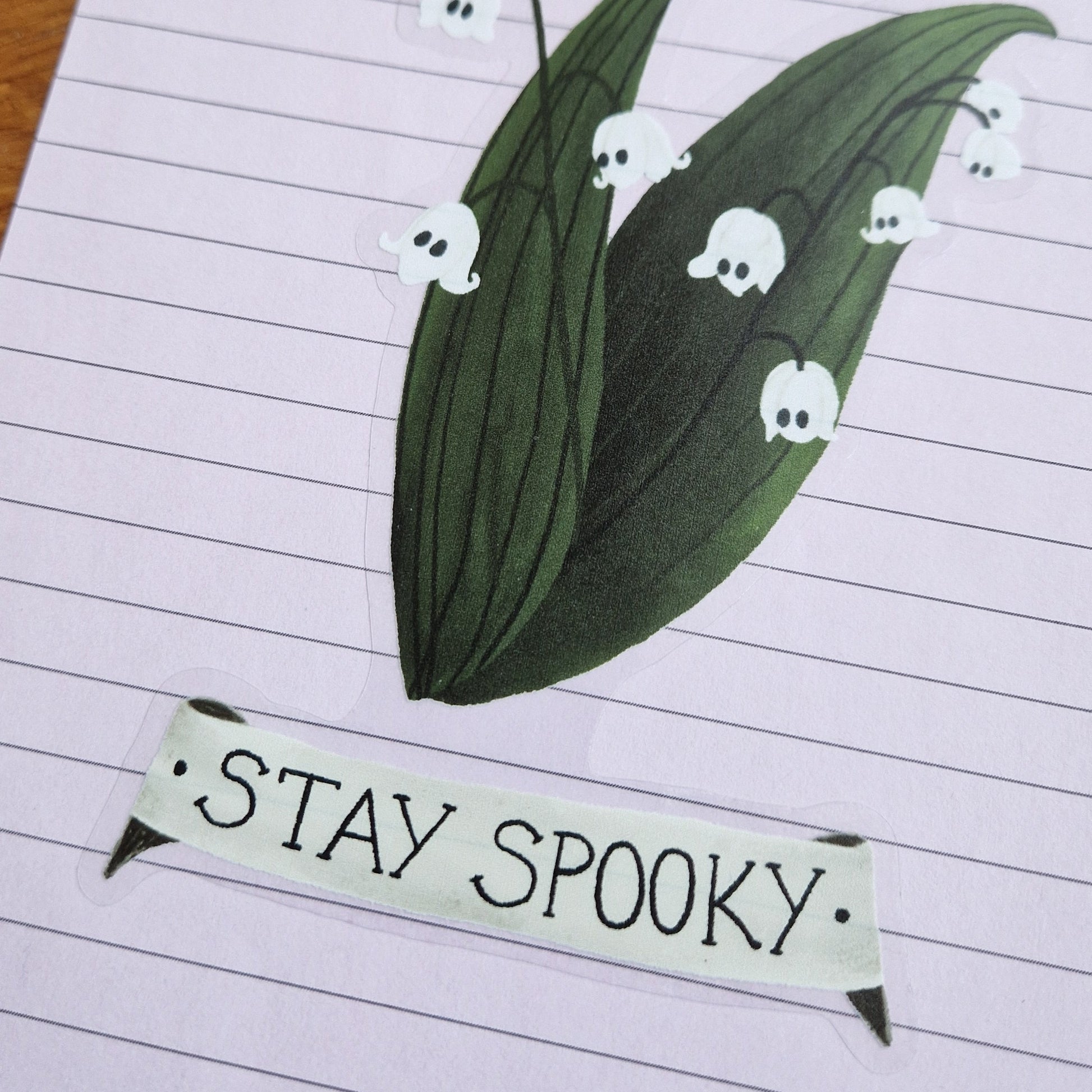 A closer view of the lined lilac piece of paper with a sticker on it. The sticker has a transparent background with a lily of the valley plant with ghost flowers and a scroll underneath with the words 'Stay Spooky'/
