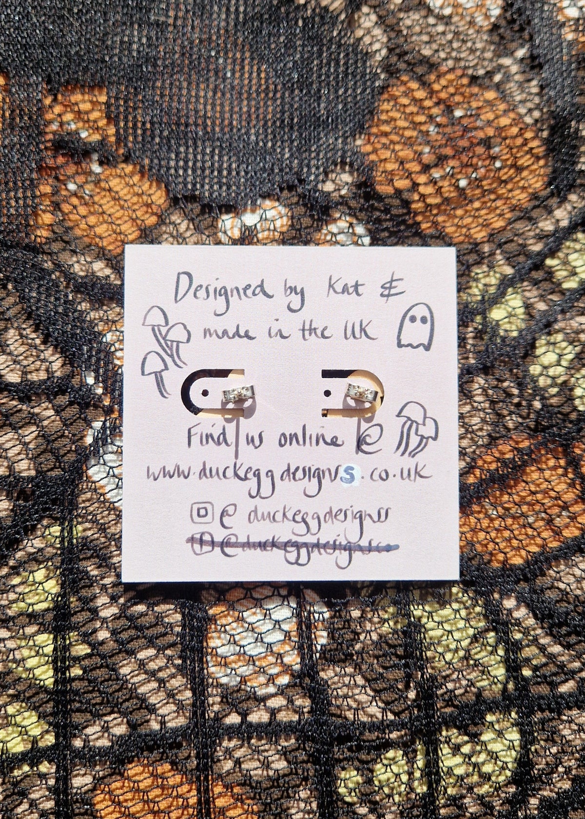 A light purple backing card sits on a black lace and floral fabric background. The backing card has a pair of silver posts and earring backs visible on it as well as black writing that reads 'Designed by Kat & made in the UK Find us online @ www.duckeggdesignss.co.uk'.