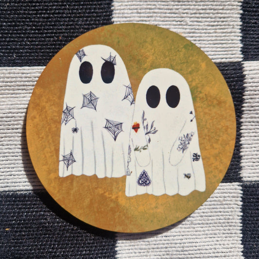 An orange round coaster with two ghosts that have tattoos. Behind the coaster you can see a checkered black and white fabric background.