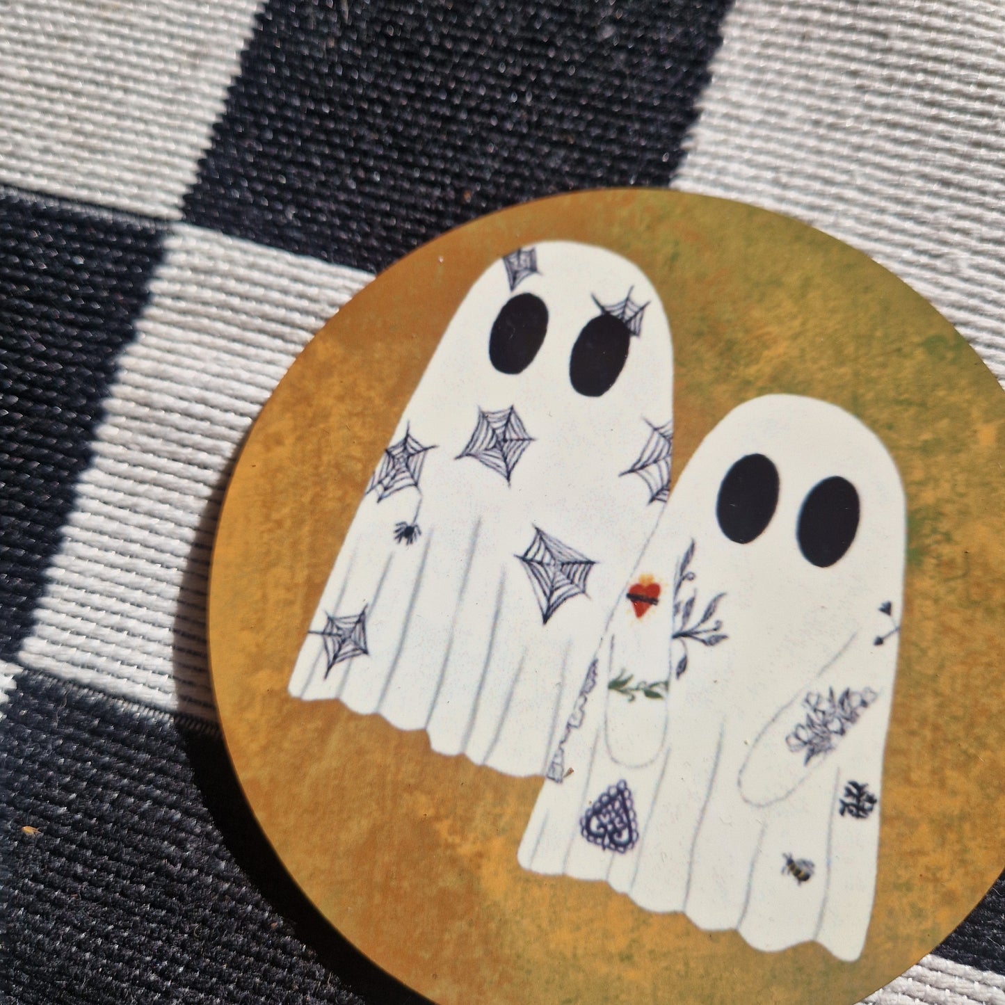 A close up of the round orange coaster showing the tattooed ghosts design up close. Behind the coaster is a black and white checkered fabric.