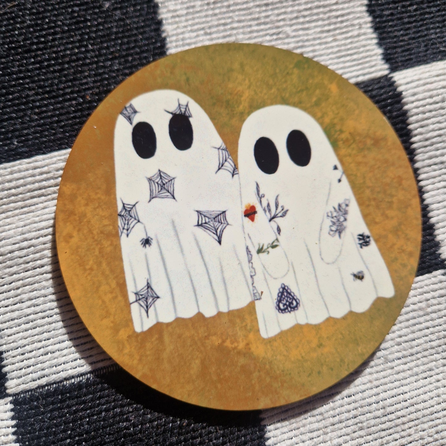 A round orange coaster sits on a checkered white and black fabric. The coaster has two ghosts which have tattoos.
