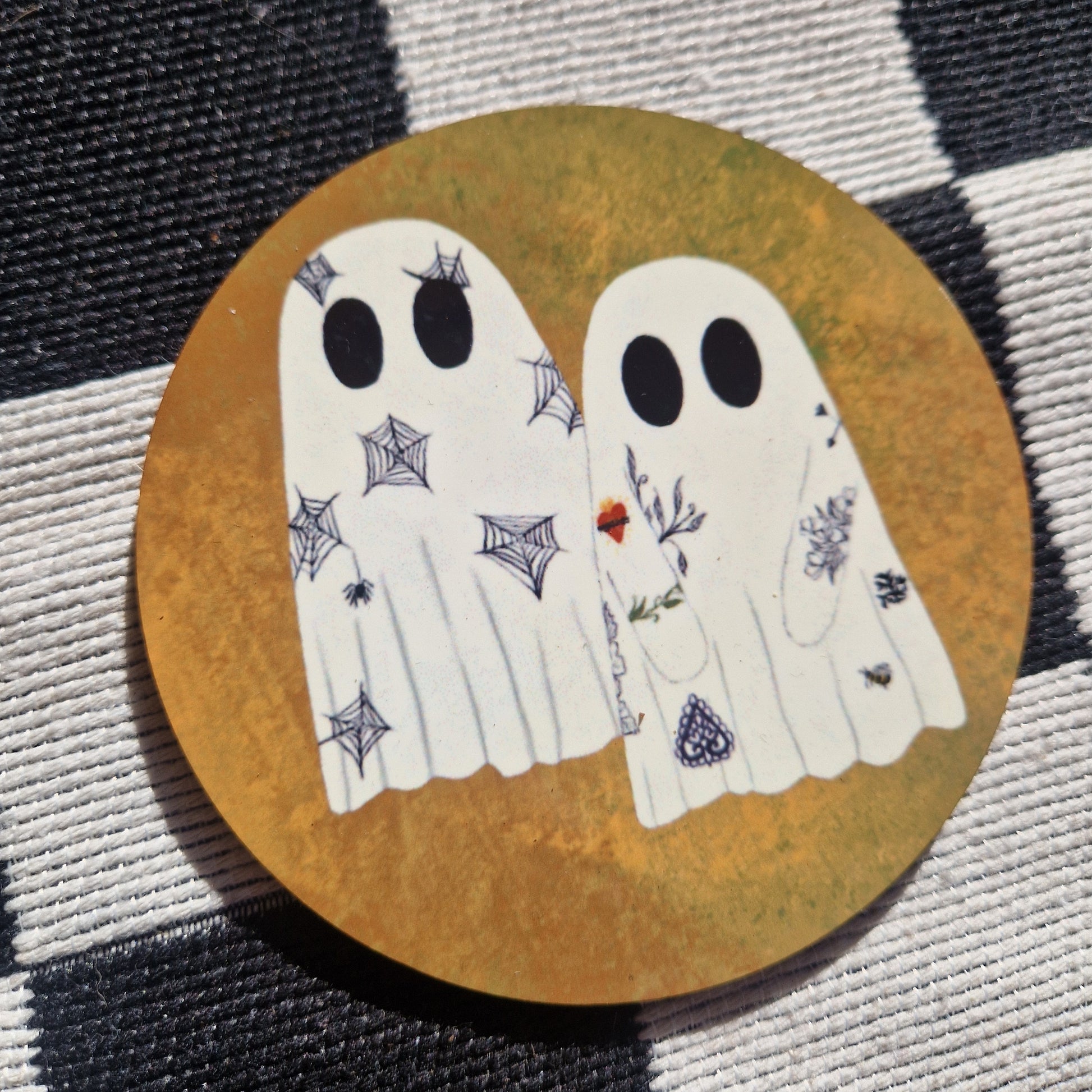 A round orange coaster sits on a checkered white and black fabric. The coaster has two ghosts which have tattoos.