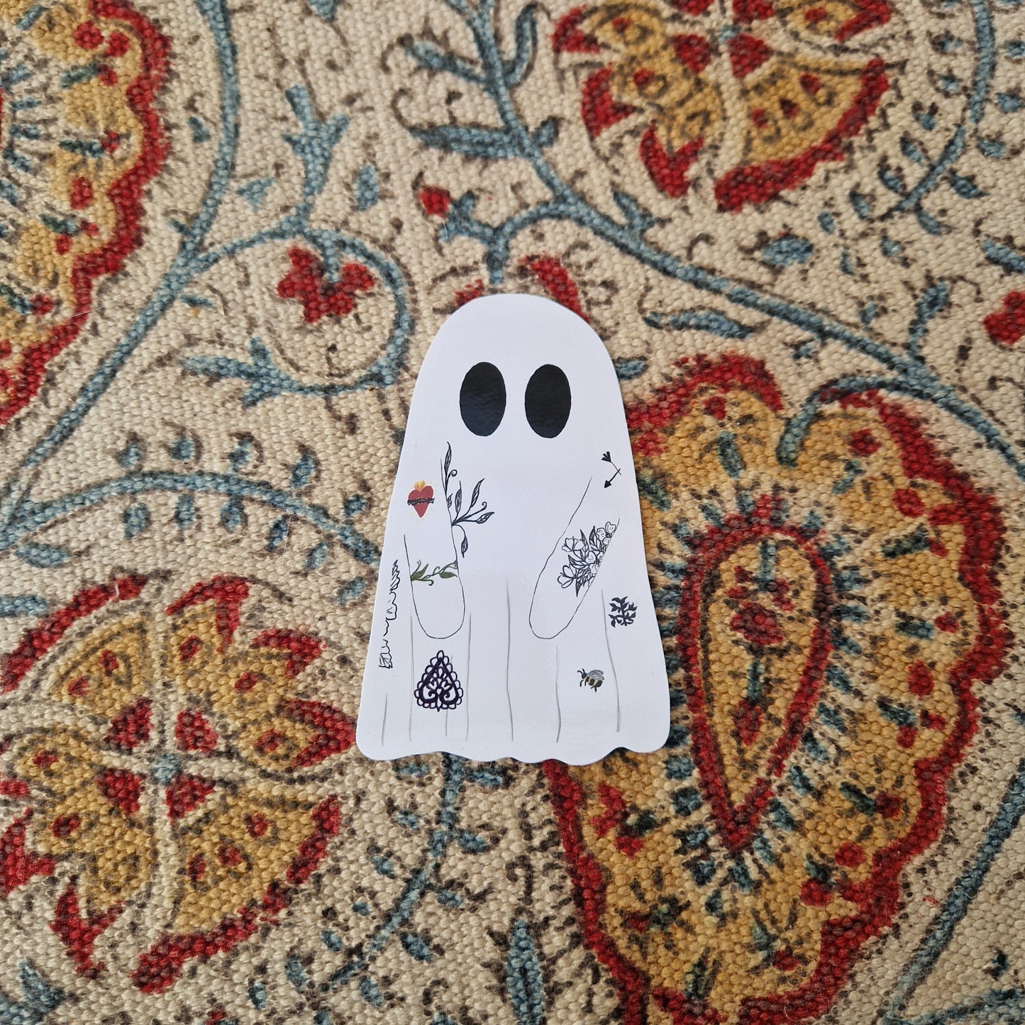 A tattooed ghost sticker sits on a patterned fabric background. The ghost has 9 different  tattoos.