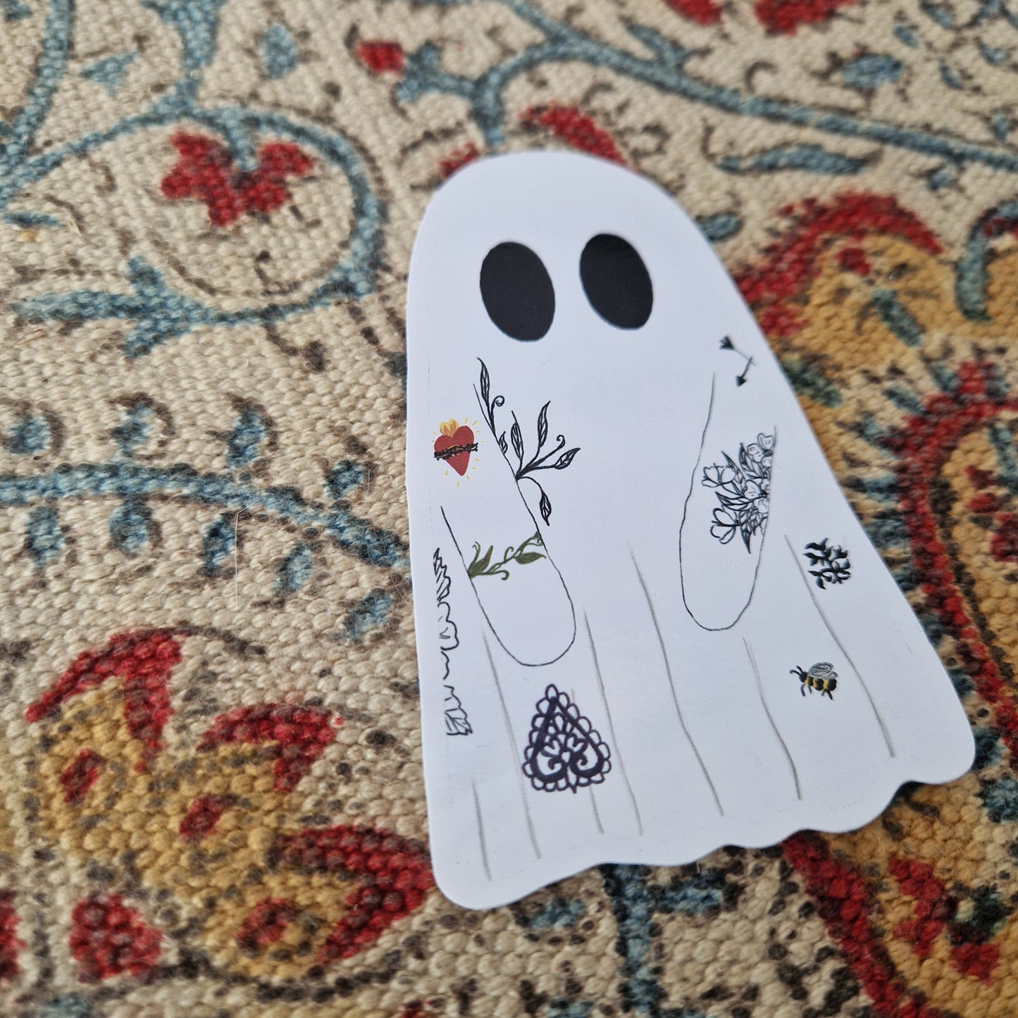 A ghost sticker with tattoos sits on a patterned fabric background.