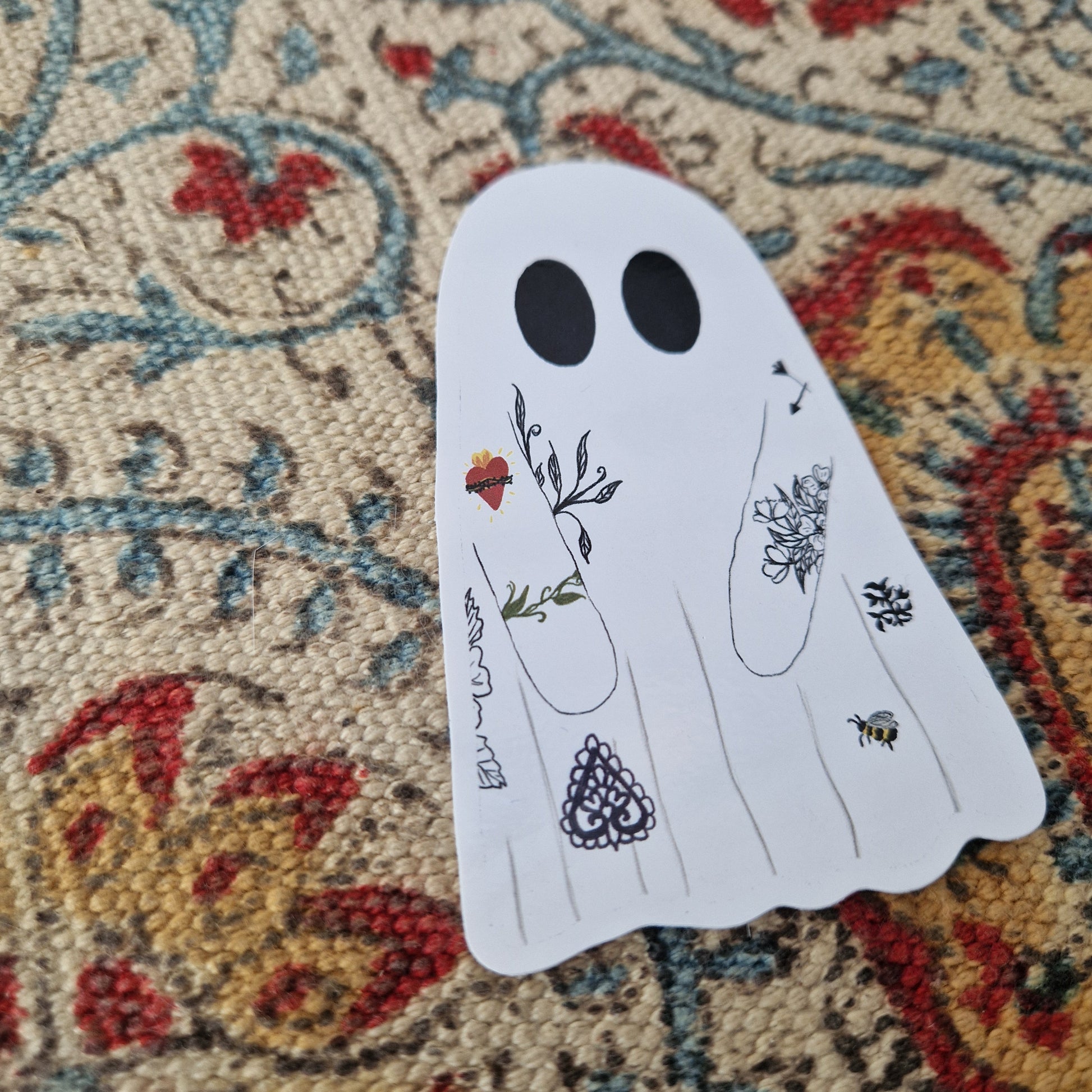 A ghost sticker with tattoos sits on a patterned fabric background.