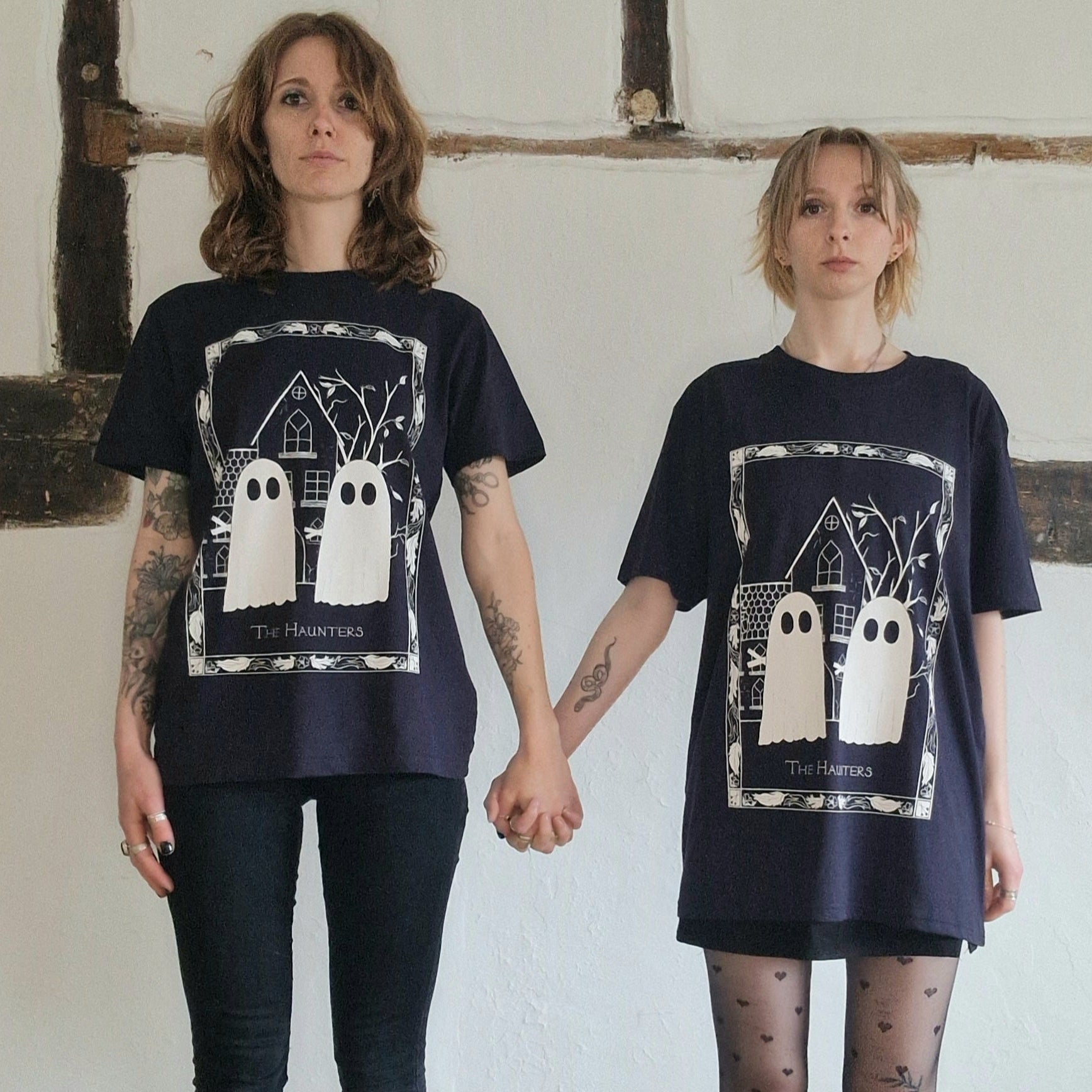 Two tattooed white women holding hands and both wearing the dark navy haunters tshirt - one with a pair of black skinny jeans and the other with a black skirt and heart patterned tights. They stand in front of a white wall with exposed wooden beams running through it.