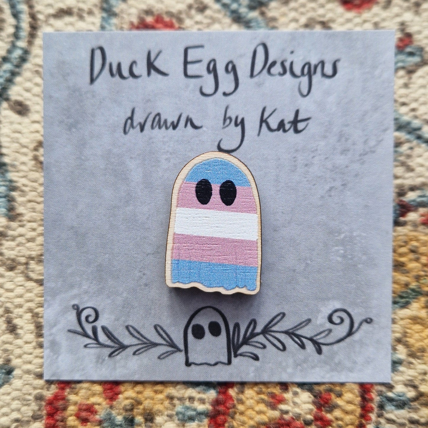 A ghost pin featuring the blue, pink and white striped design of the transgender flag. The ghost is on a grey backing card with the words 'Duck Egg Designs drawn by Kat' in black handwriting. Behind the backing card is a textured fabric background.