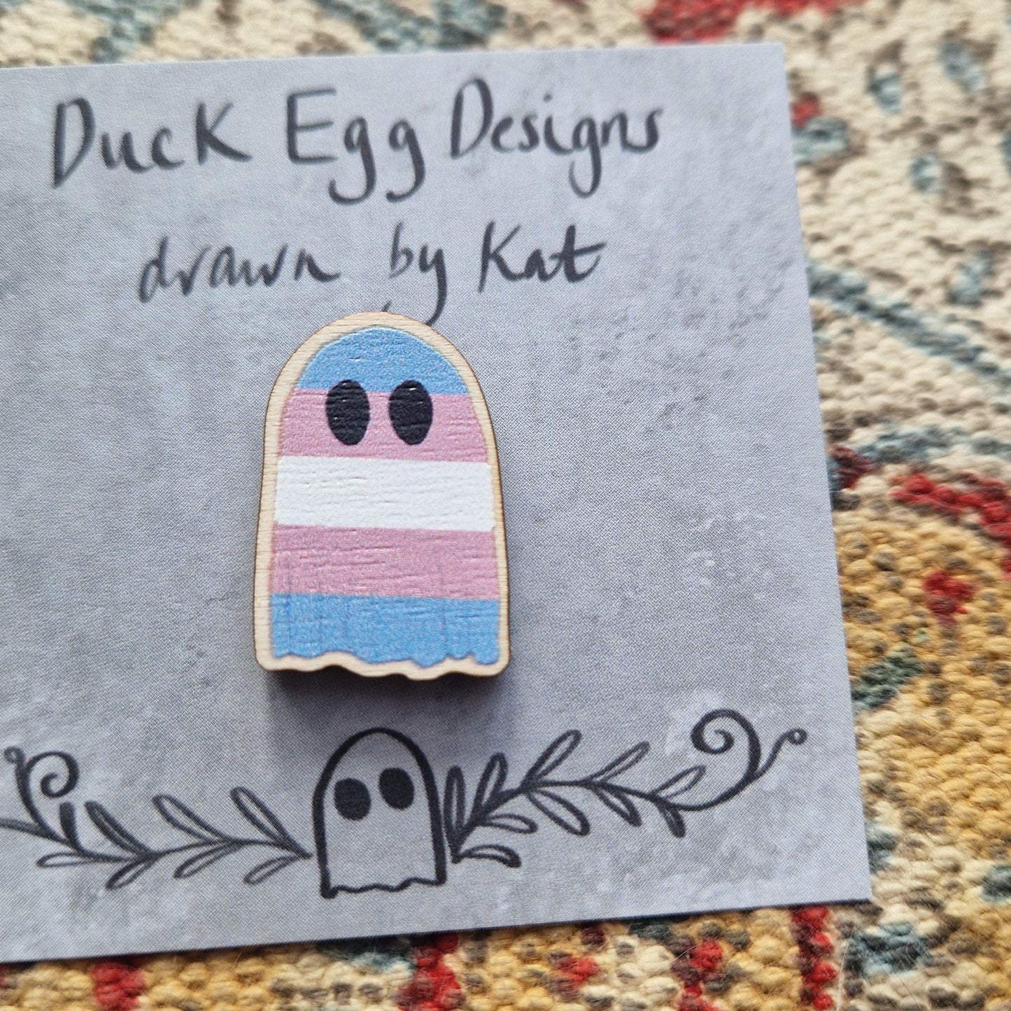 A ghost pin featuring the blue, pink and white striped design of the transgender flag. The ghost is on a grey backing card with the words 'Duck Egg Designs drawn by Kat' in black handwriting. Behind the backing card is a textured fabric background.