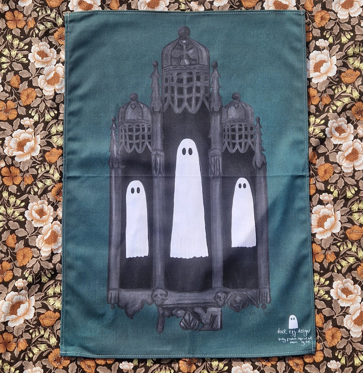 A dusty blue tea towel featuring grey gothic architecture and three floating ghosts sits on a floral fabric background,