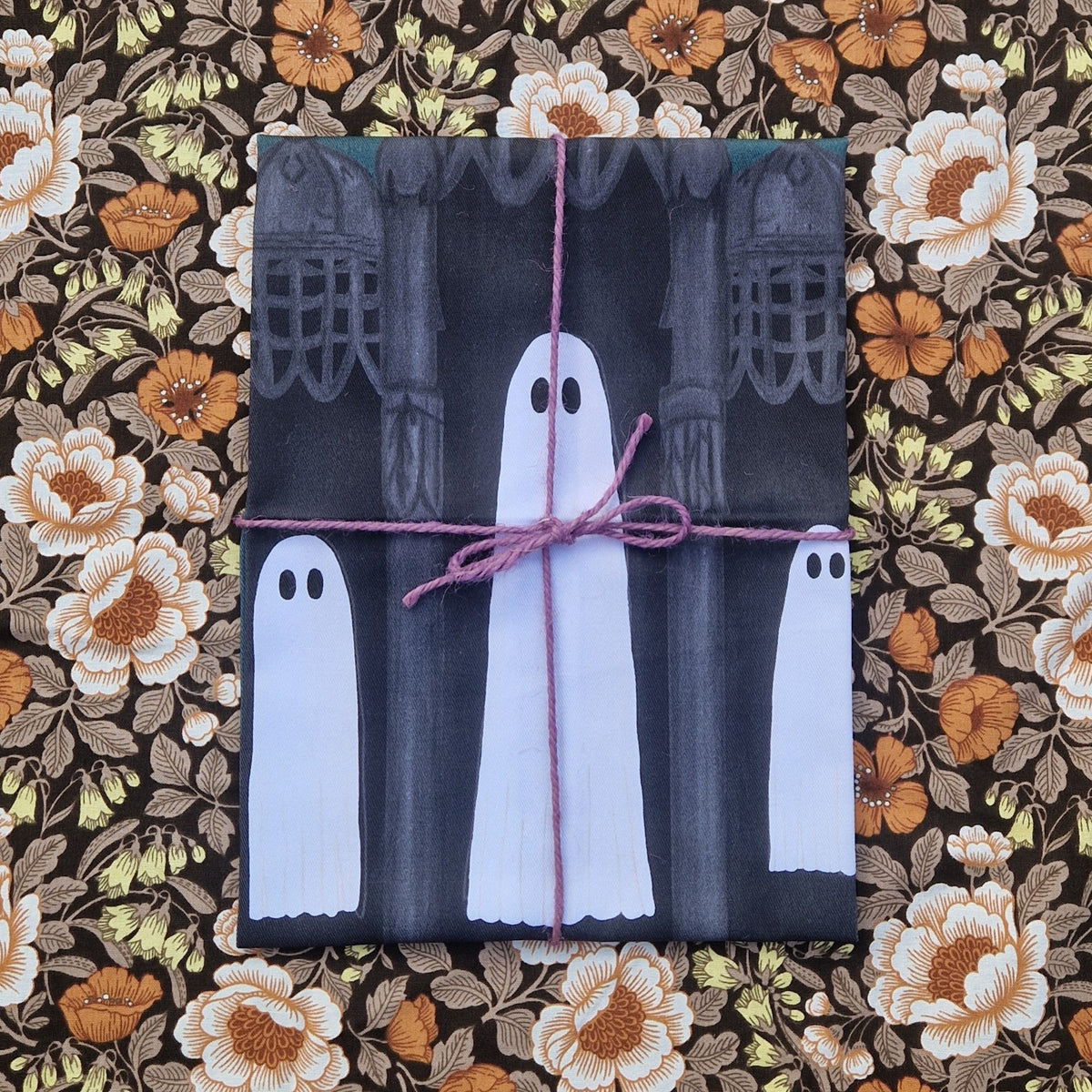 A  folded teatowel sits on a floral patterned background wrapped in a purple twine. Visible on the teatowel are three ghosts floating next to each other surrounded by gothic architecture.