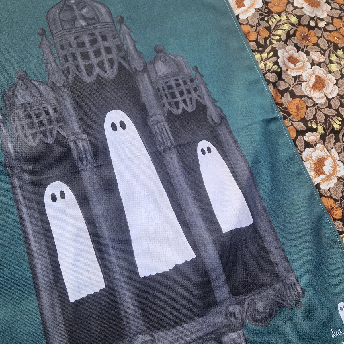 A close up view of the dusty blue teatowel showing the details in the ghost illustration and the architecture they float in. To the right of the teatowel you can see a floral patterned background.