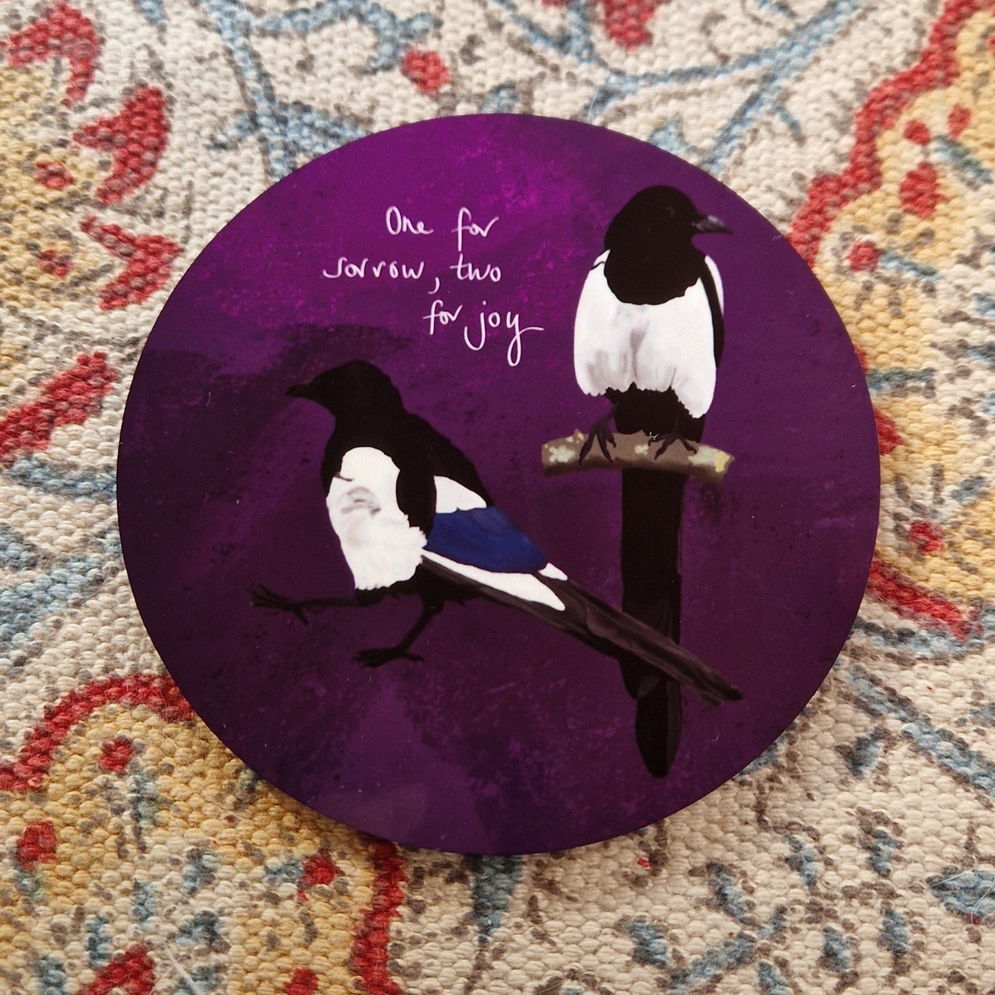 A round deep purple coaster with two magpies and the words 'One for sorrow, tow for joy' in white handwriting. The coaster sits on a patterned fabric background.