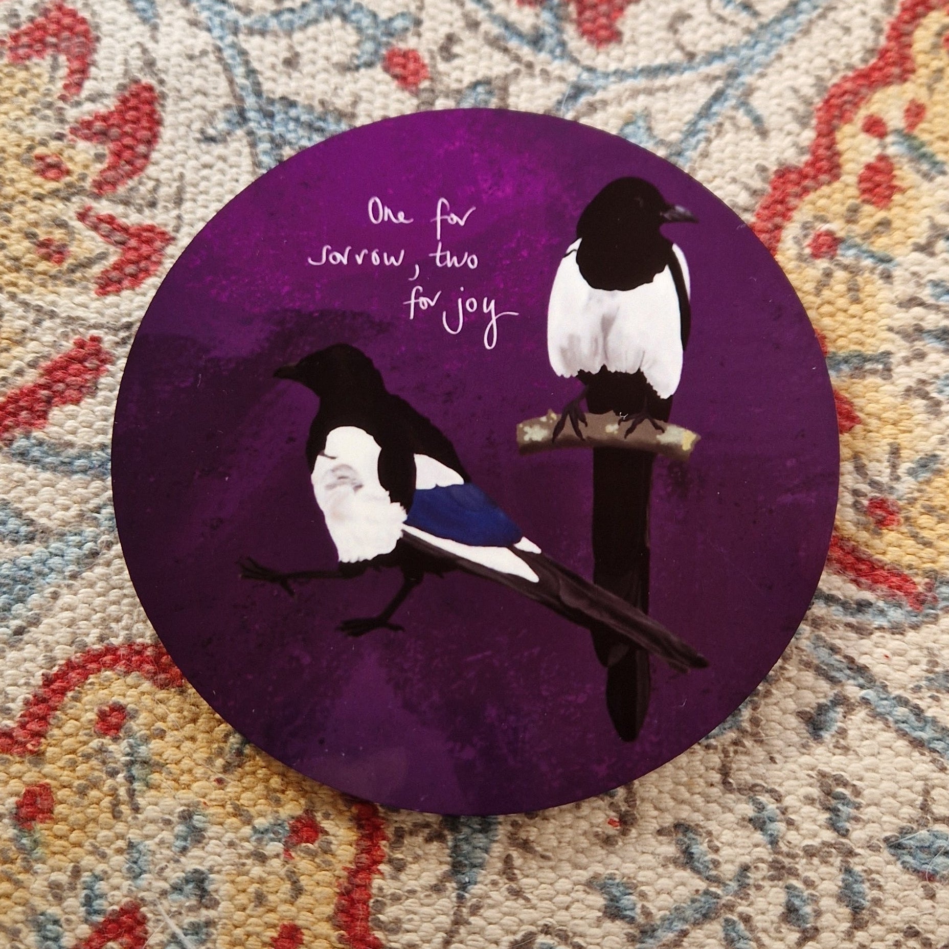 A round deep purple coaster with two magpies and the words 'One for sorrow, tow for joy' in white handwriting. The coaster sits on a patterned fabric background.