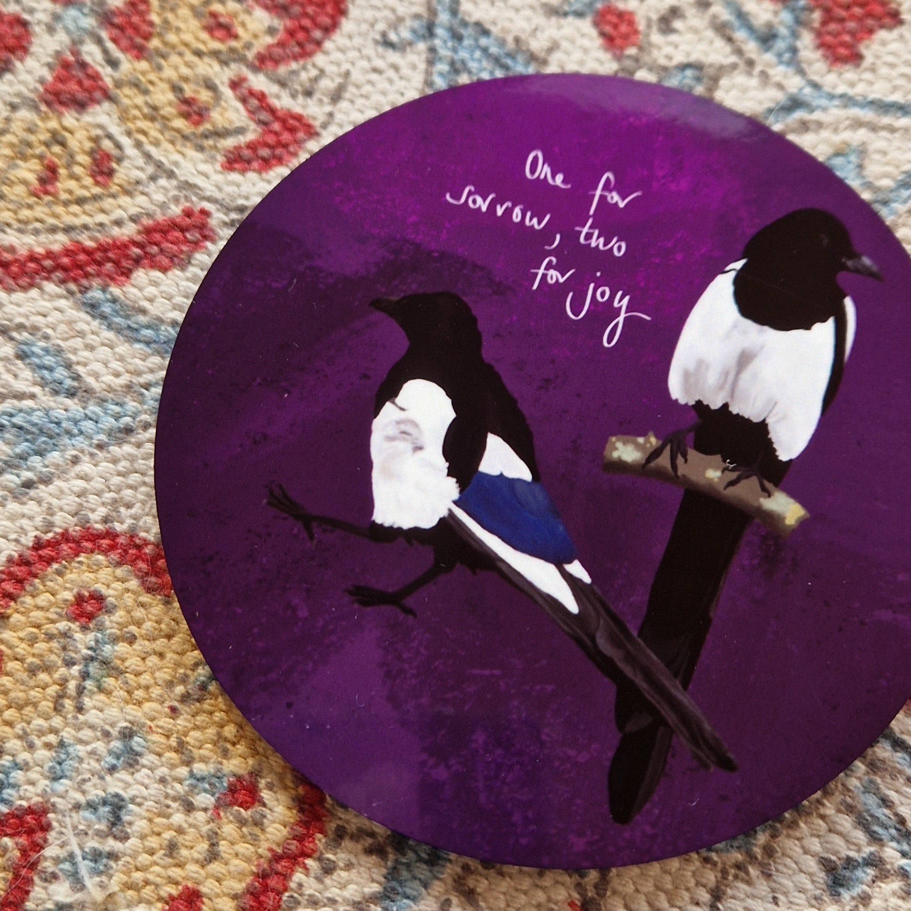 A close up of the purple coaster showing the detail in the two magpies illustration. The coaster sits on a patterned fabric background.