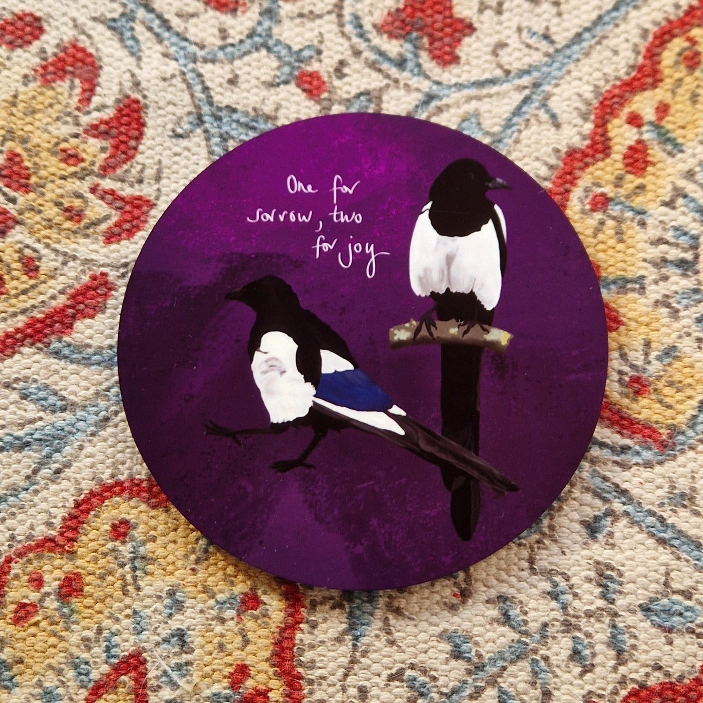 A round deep purple coaster with two magpies and the words 'One for sorrow, tow for joy' in white handwriting. The coaster sits on a patterned fabric background.