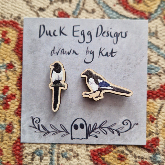 Two small magpie pin badges sit on a grey backing card with the words 'Duck Egg Designs drawn by Kat' across the top. Behind the backing card is a patterned fabric background.
