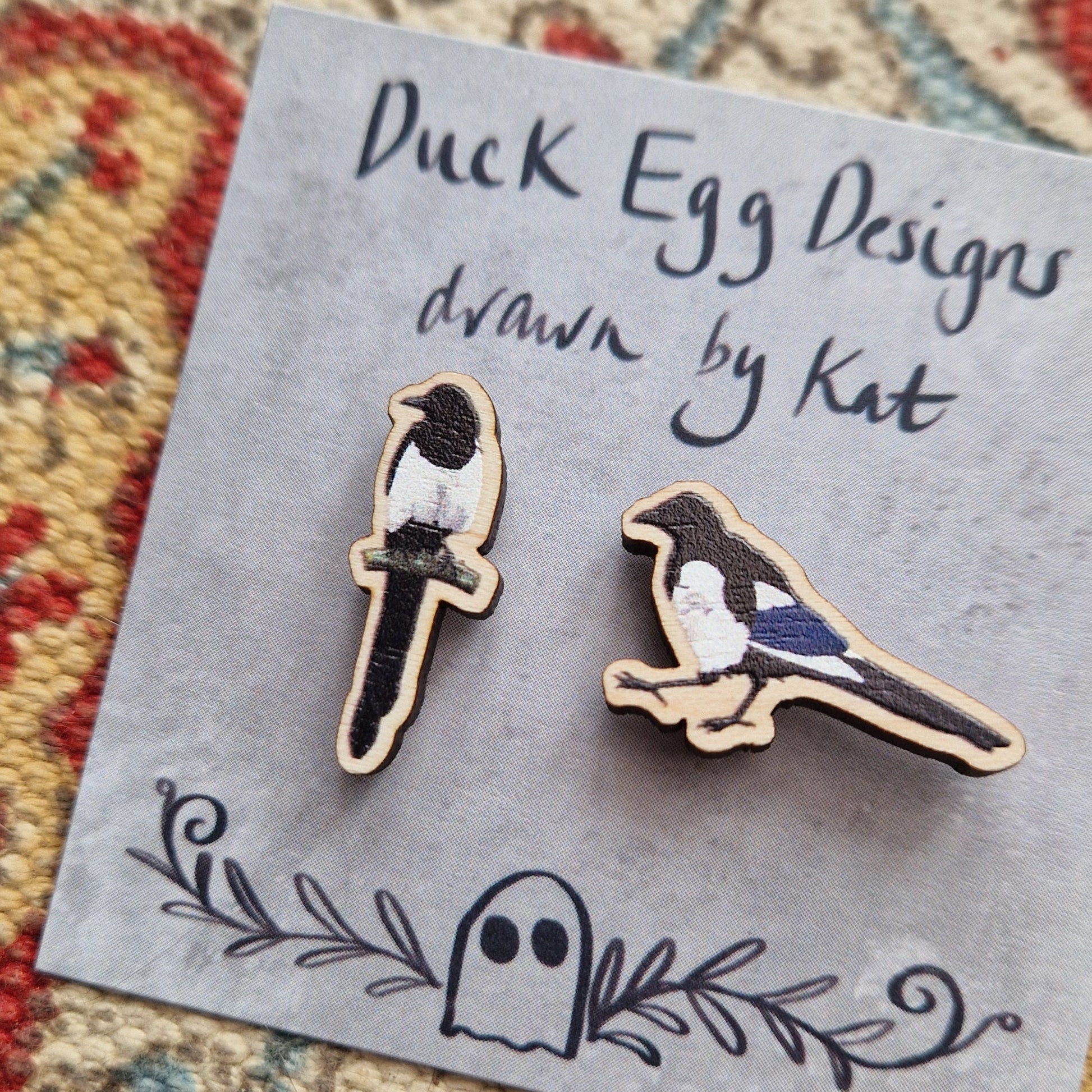 A close up of the two magpie pin badges, demonstrating the texture of the wooden pins. The pins sit on a grey backing card with the words 'Duck Egg Designs drawn by Kat' in black handwriting across the top. Behind the backing card you can see a patterned fabric background.