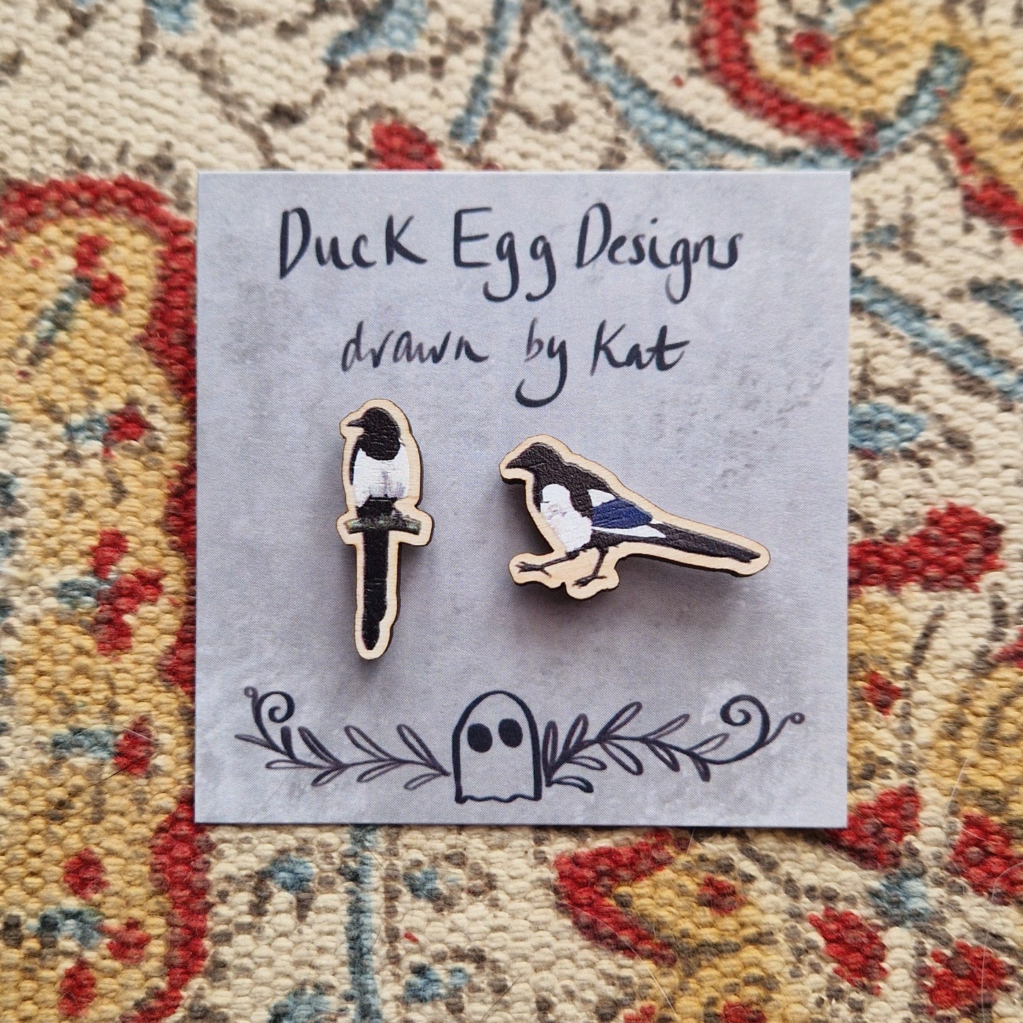 Two small magpie pin badges sit on a grey backing card with the words 'Duck Egg Designs drawn by Kat' across the top. Behind the backing card is a patterned fabric background.