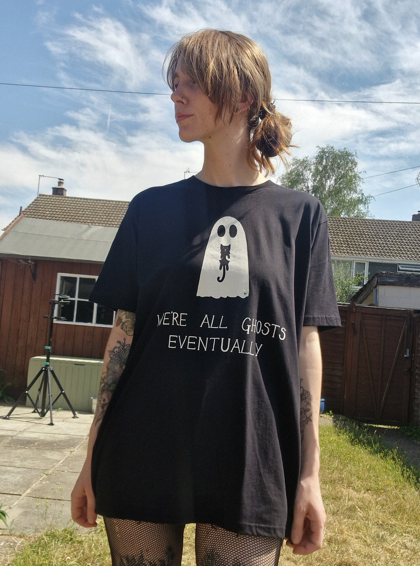 A white woman with ginger hair wears an oversized black t-shirt featuring a white ghost holding a black cat with the phrase 'We're all ghosts eventually' written underneath. Behind the person is a sunny garden with gteen grass.