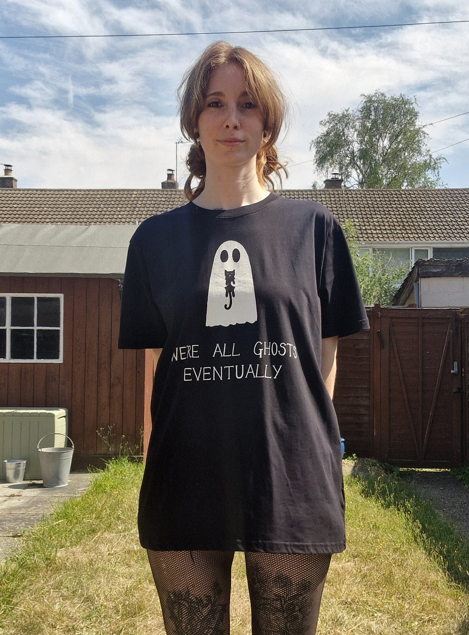 A white woman with ginger hair wears an oversized black t-shirt featuring a white ghost holding a black cat with the phrase 'We're all ghosts eventually' written underneath. Behind the person is a sunny garden with gteen grass.