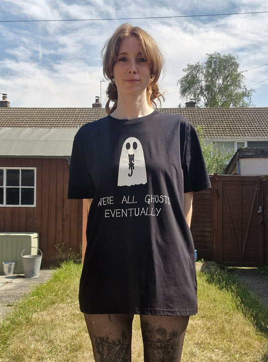 A white woman with ginger hair wears an oversized black t-shirt featuring a white ghost holding a black cat with the phrase 'We're all ghosts eventually' written underneath. Behind the person is a sunny garden with gteen grass.
