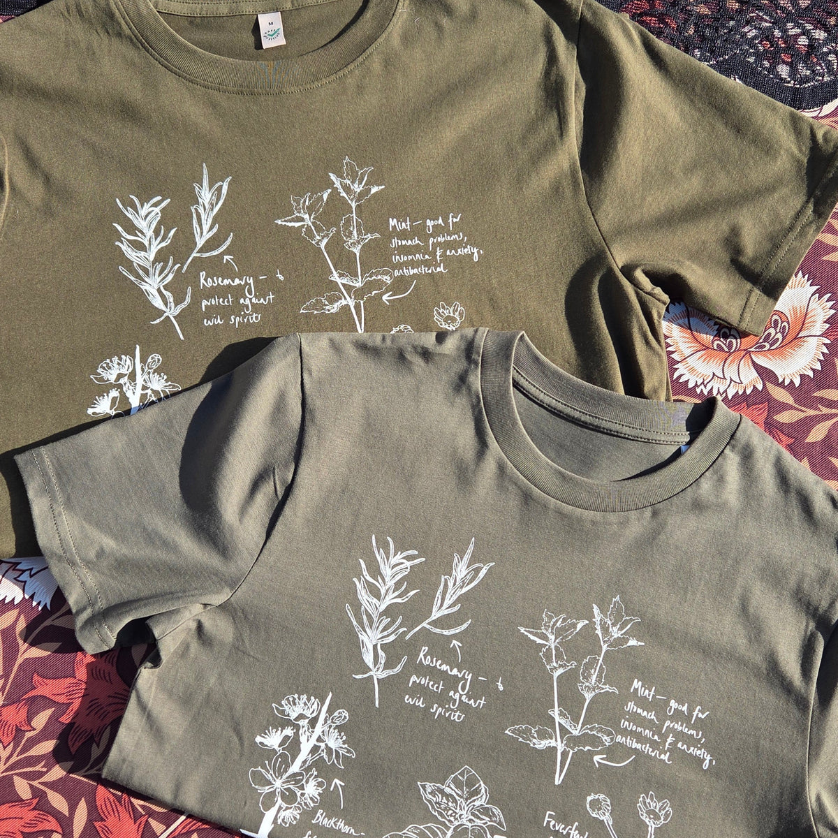 Two folded t-shirts laid side by side one sage green and the other mossy green, both with a plant filled design in white on them. 