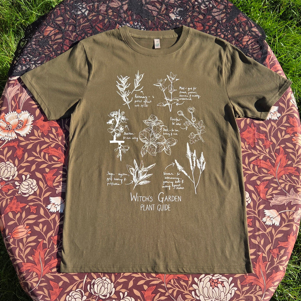 A mossy green shirt with a white plant filled design on the front above the words 'Witch's Garden Plant Guide' behind the t-shirt is a floral brown patterned fabric with a black lace overlay.