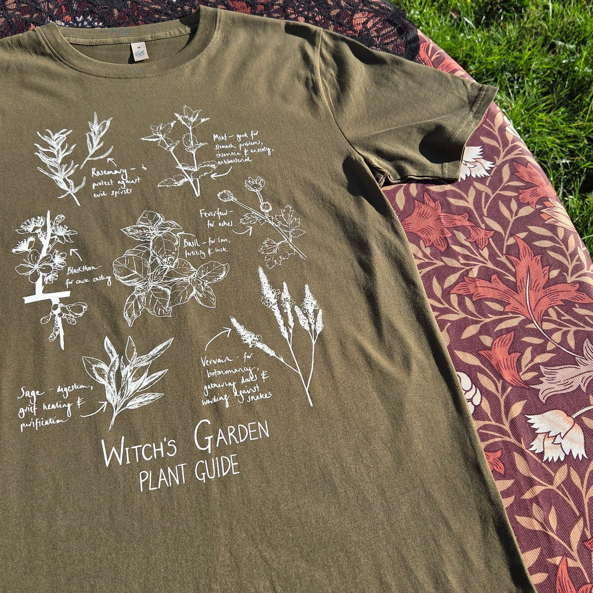 A close up of a mossy green shirt with a white plant filled design on the front above the words 'Witch's Garden Plant Guide' behind the t-shirt is a floral brown patterned fabric with a black lace overlay.