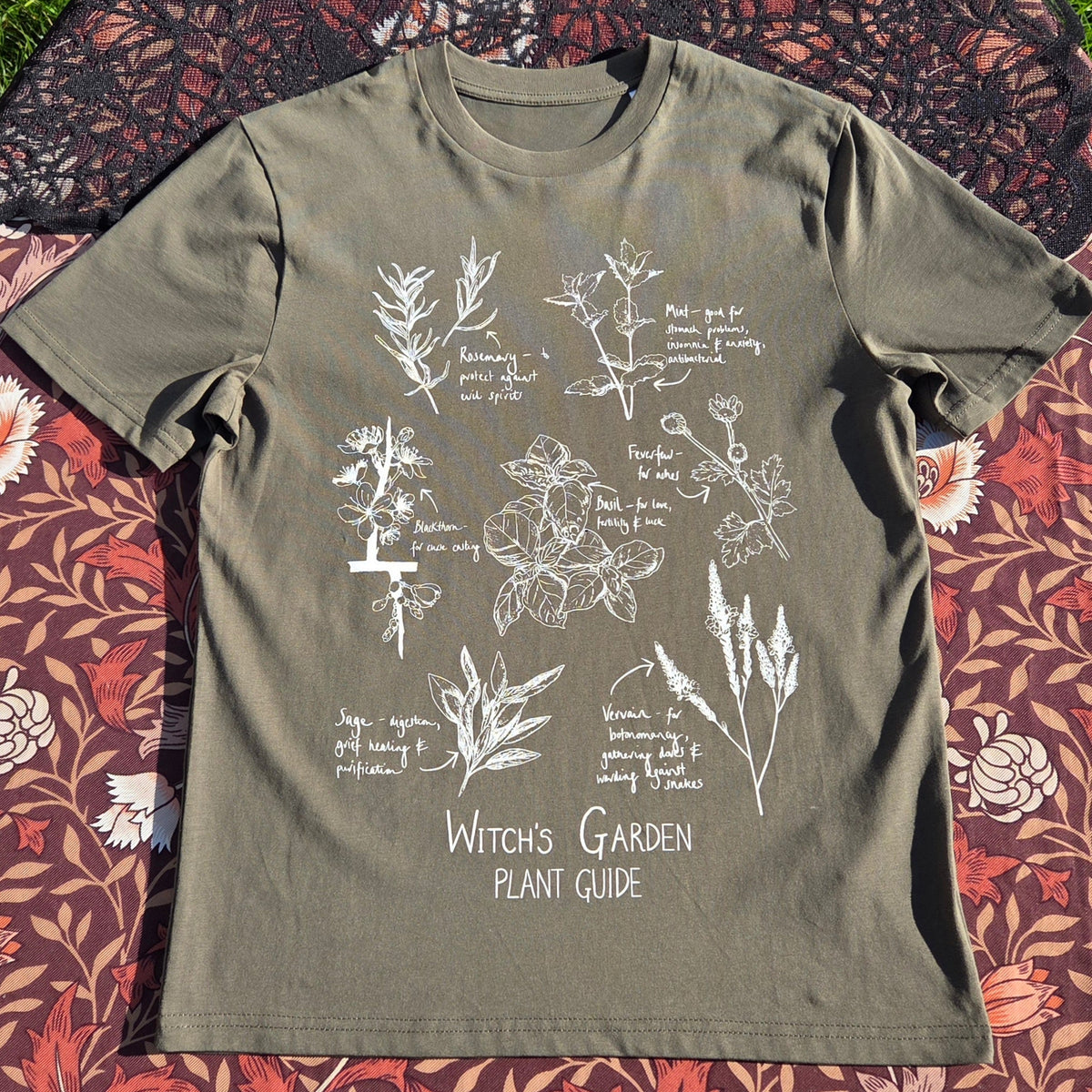 A sage green shirt with a white plant filled design on the front above the words 'Witch's Garden Plant Guide' behind the t-shirt is a floral brown patterned fabric with a black lace overlay.