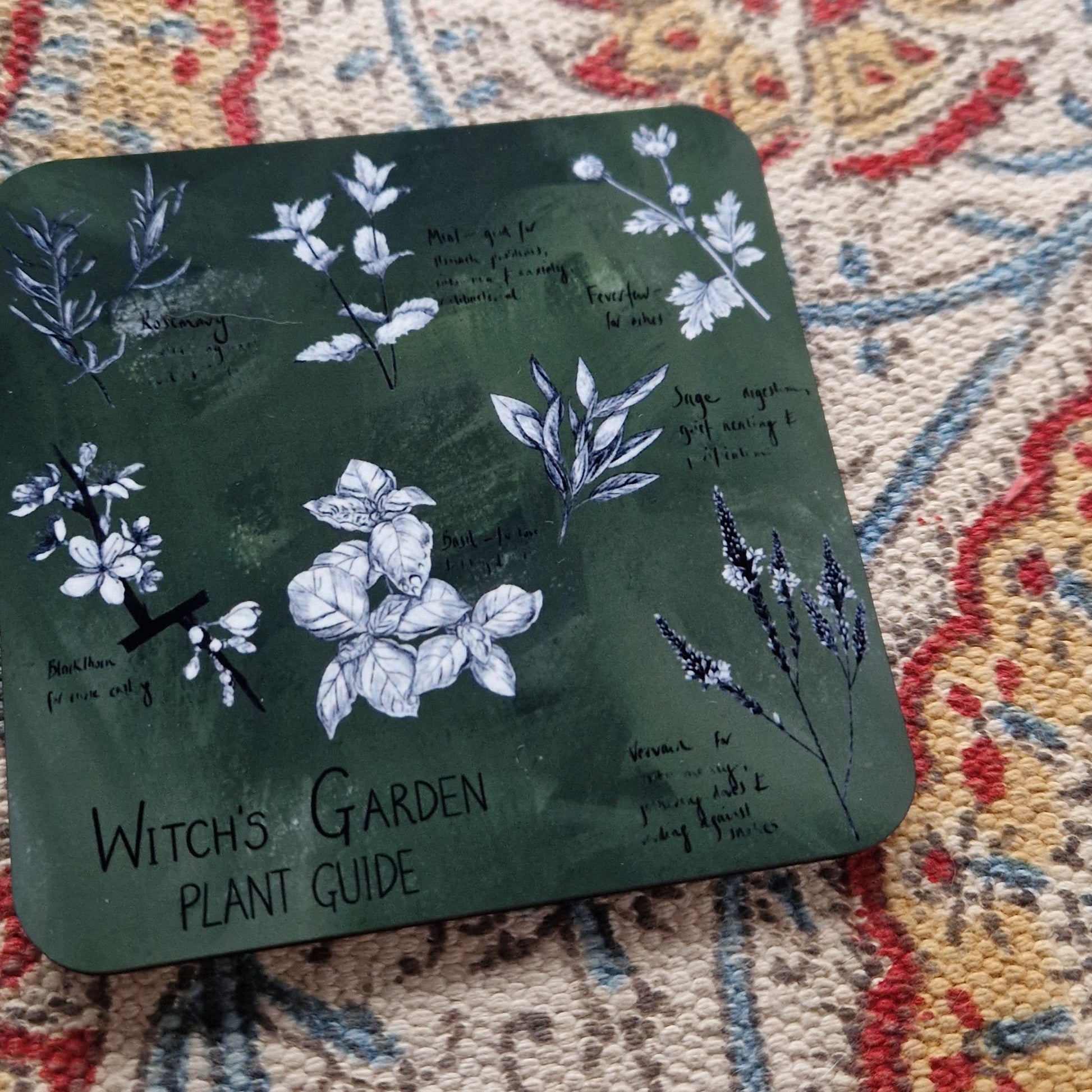 A closer look at the green square coaster featuring seven different plant illustrations and the words 'Witch's Garden Plant Guide' in black handwriting.