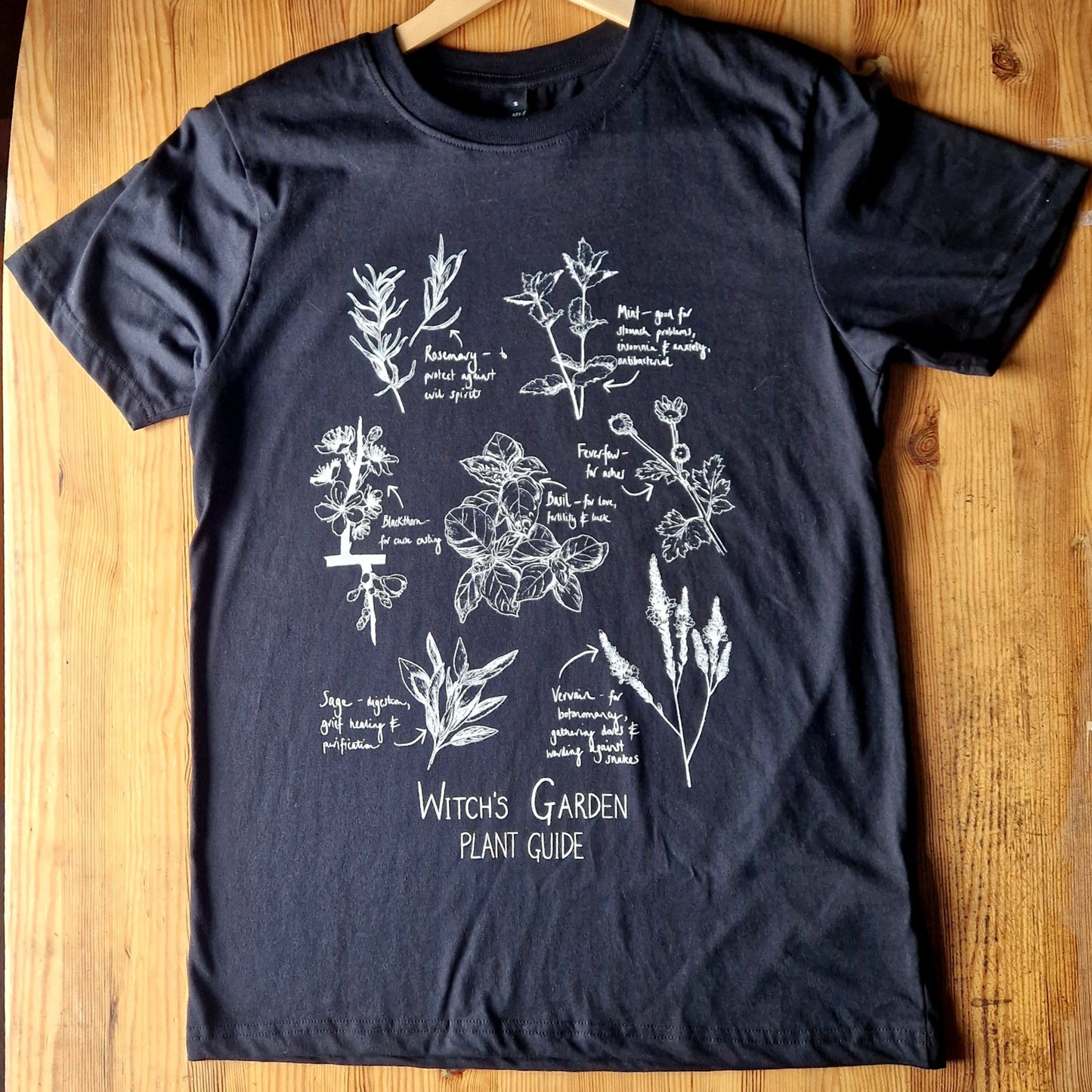 A black t-shirt lies on a wooden table. The tshirt has the words 'Witch's Garden Plant Guide' in white handwriting beneath white botanical illustrations of different plants with their names and uses next to them.