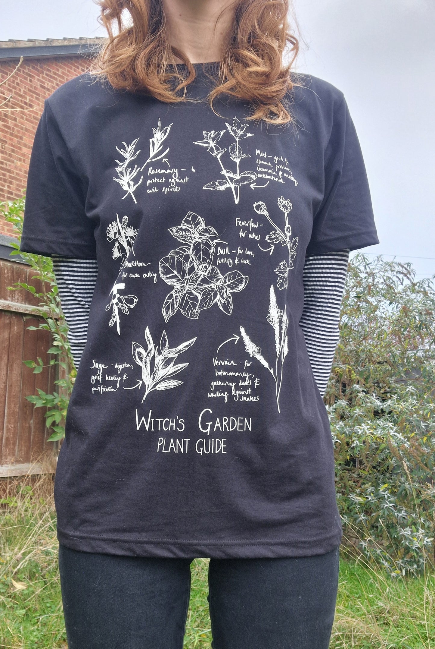 A person with long ginger hair wears the black Witch's Garden Plant Guide t-shirt with a white and black striped top underneath and black jeans. The t-shirt features white botanical plant illustrations above the words 'Witch's Garden Plant Guide'. Behind the person is an overgrown green garden.